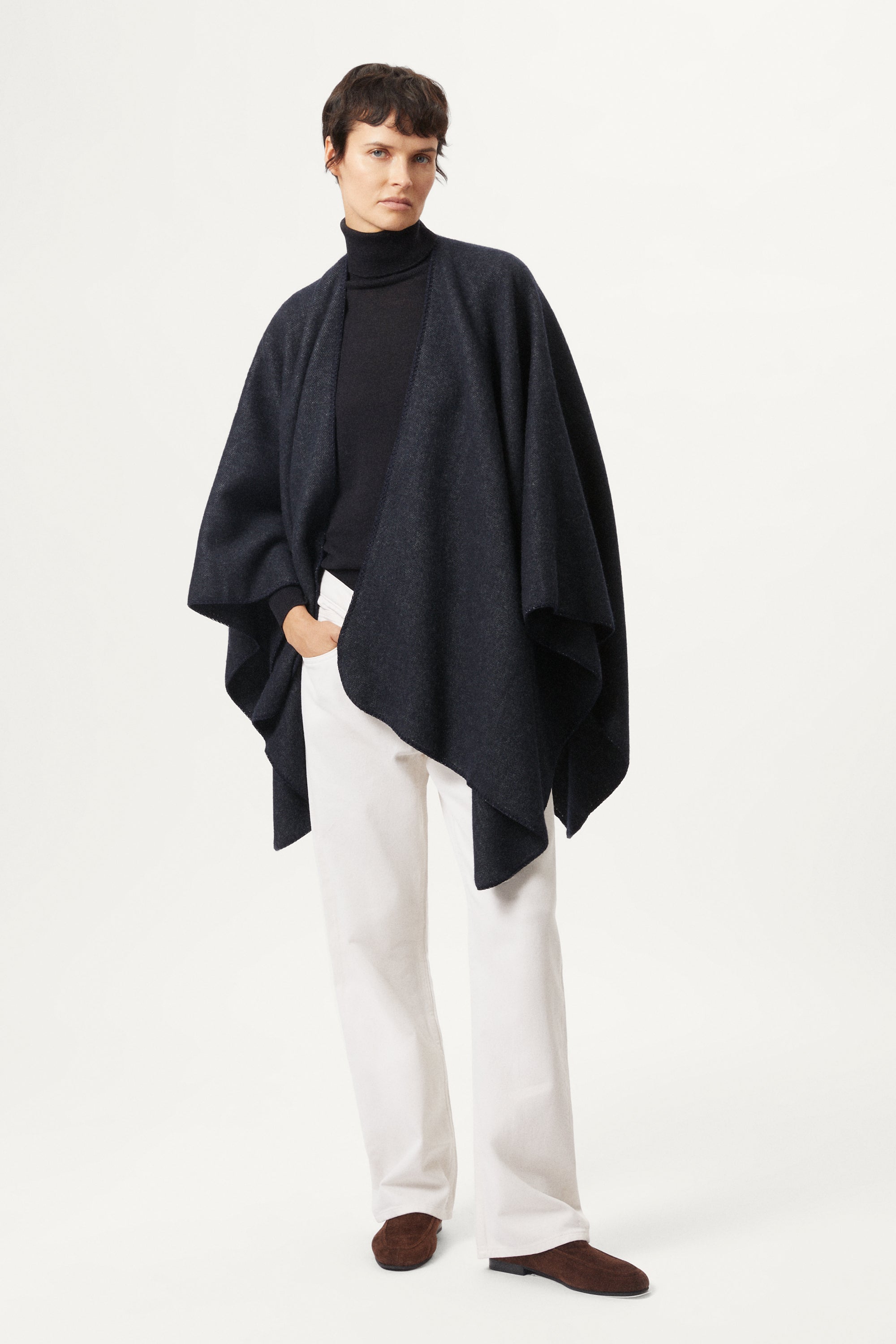 woman wearing a blue navy poncho over a turtleneck and white pants on a white background