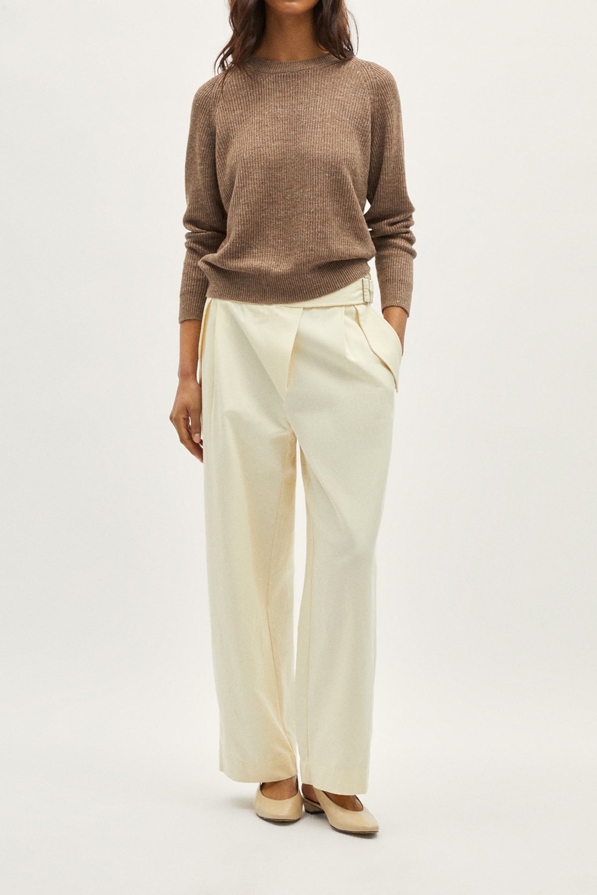 Person wearing a brown sweater and white pants on a light gray background
