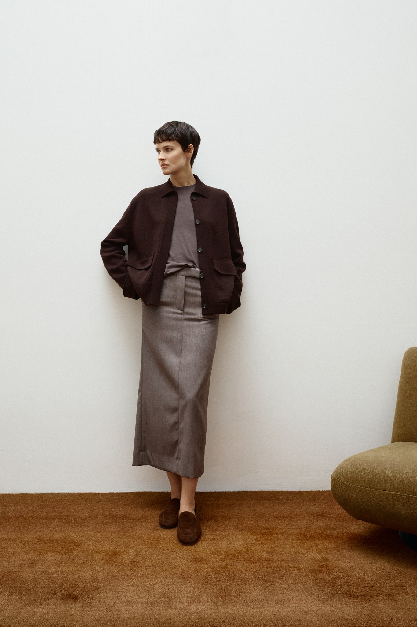 Person wearing a brown wool jacket and gray skirt standing against a white wall with a brown carpet.