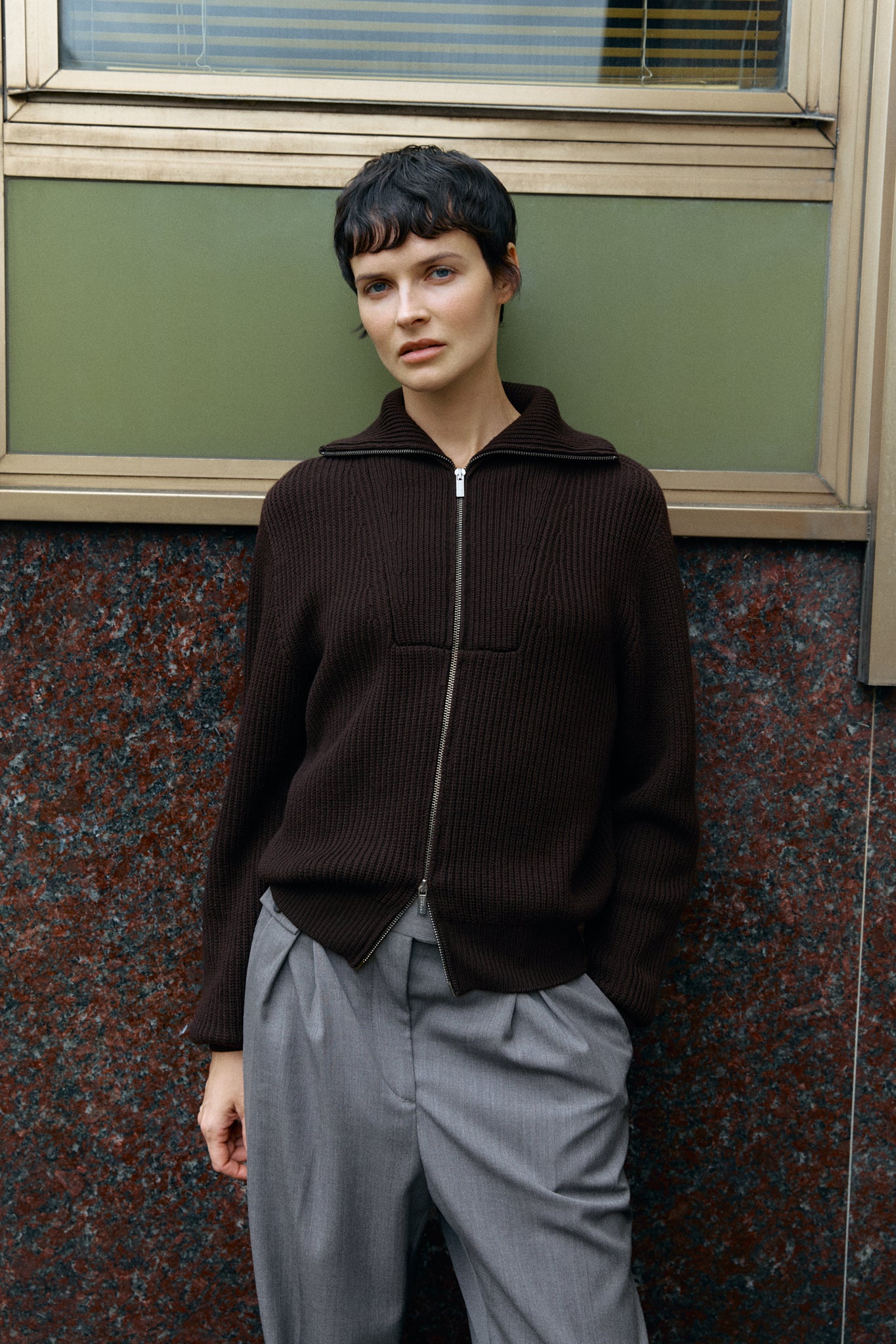 woman wearing a dark brown full zip sweater and gray pants standing against a textured wall.
