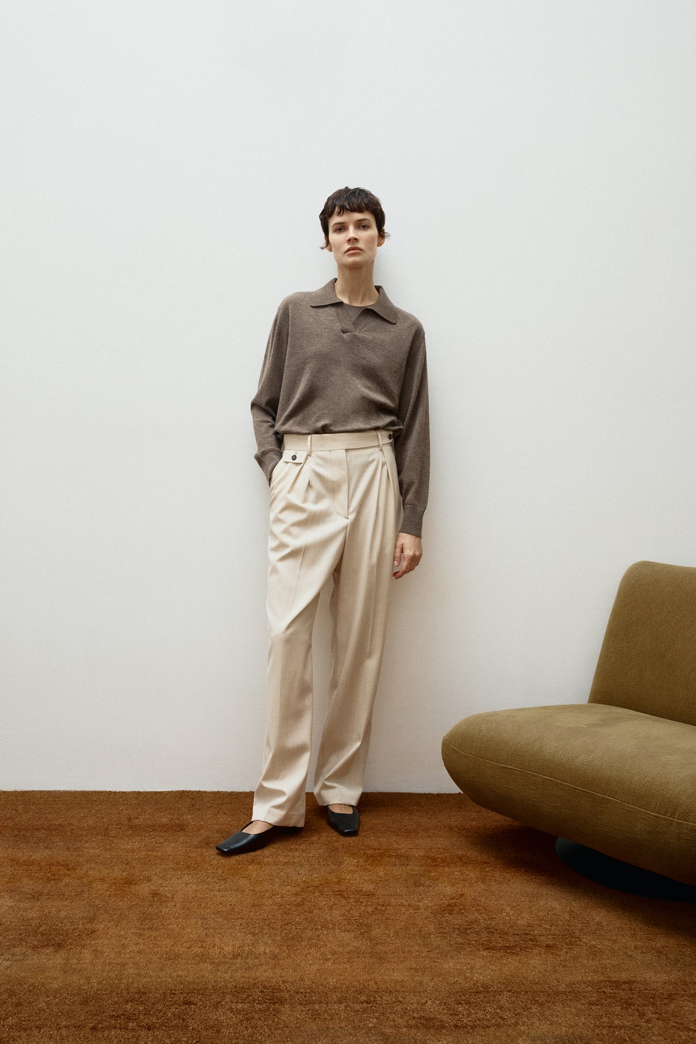 Person wearing a taupe wool polo and beige pants standing in a minimalistic room with a brown sofa.