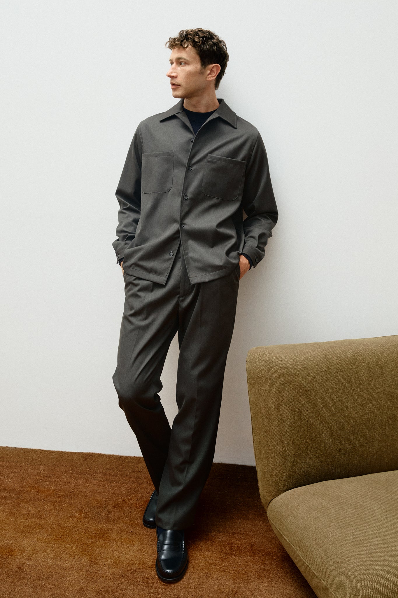 Man wearing a green outfit standing against a white wall with a brown couch.