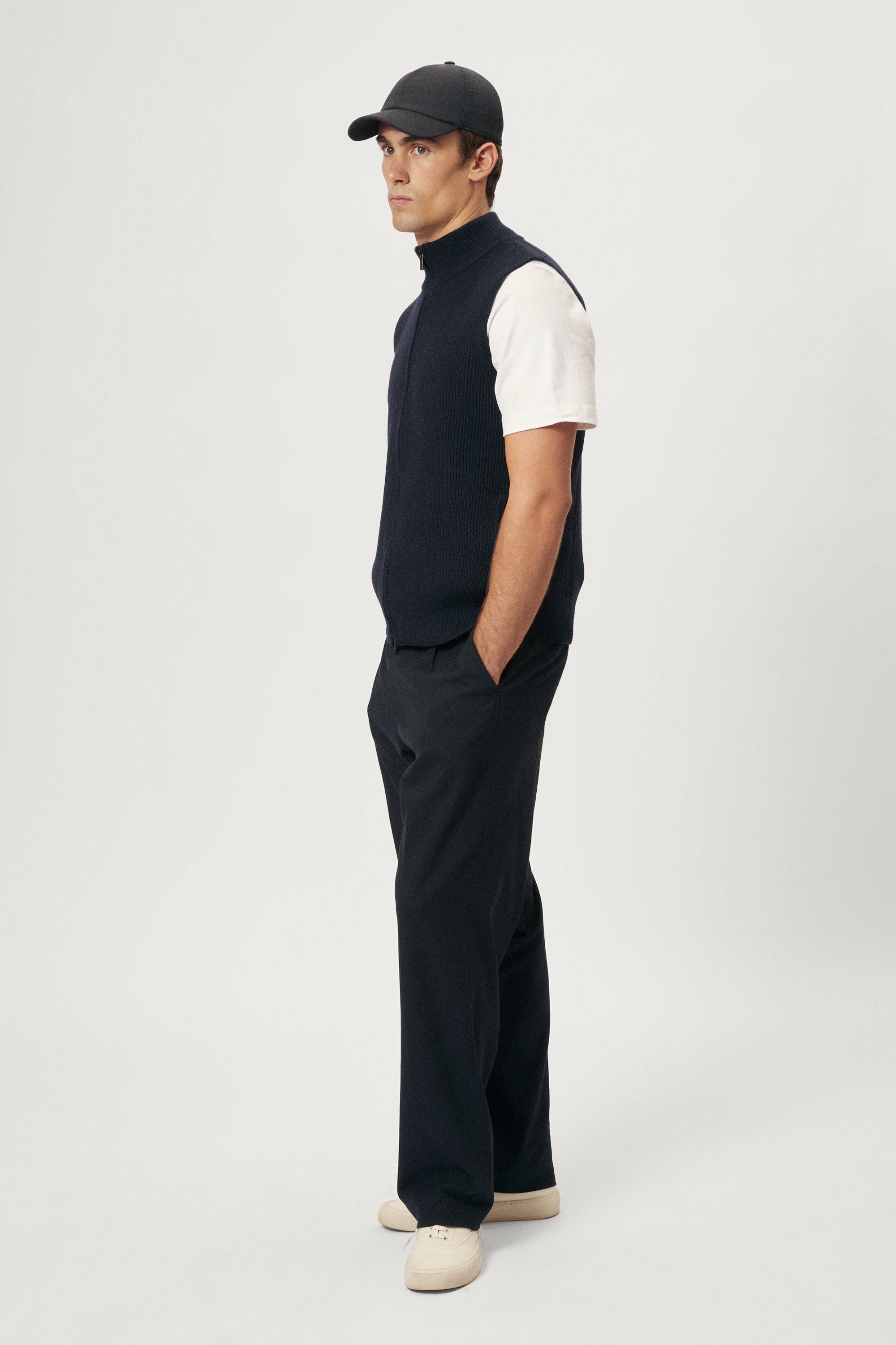 Man wearing a black vest, white shirt, black pants, and hat on a white background