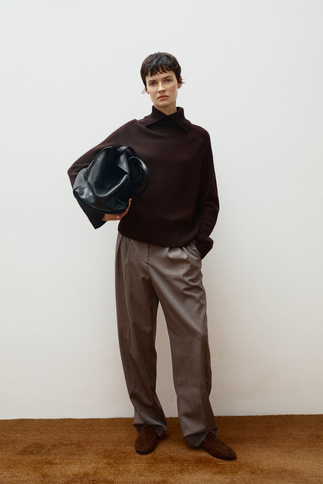 Person wearing a brown turtleneck and taupe pants holding a black leather bag against a white background