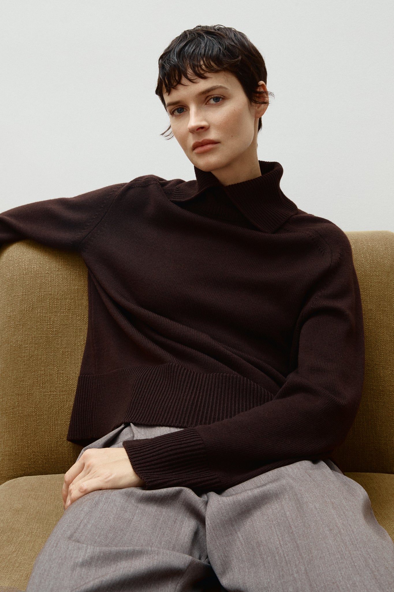Person wearing a brown Merino Wool Boxy with special High Neck on a beige couch.