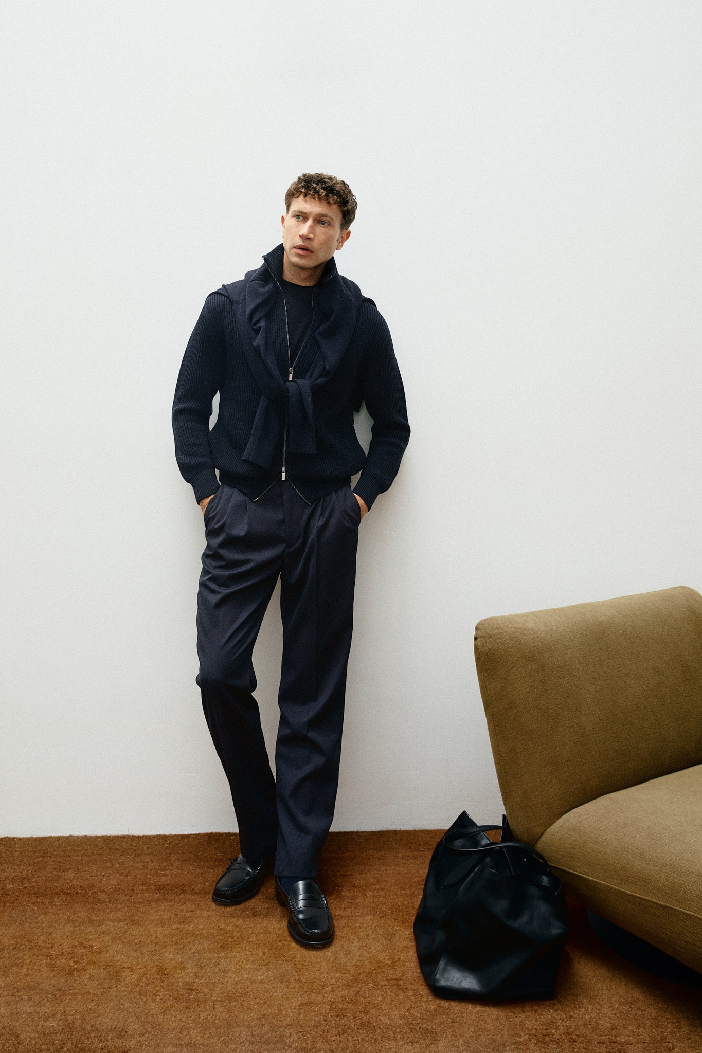 Man in a dark blue outfit standing against a white wall with a brown couch.