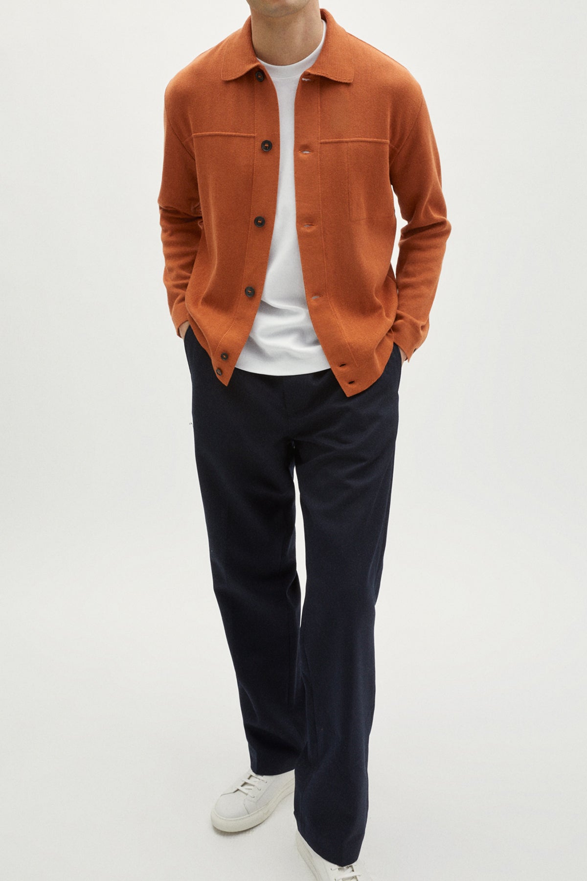 Person wearing an orange jacket, white shirt, dark pants, and white shoes on a white background