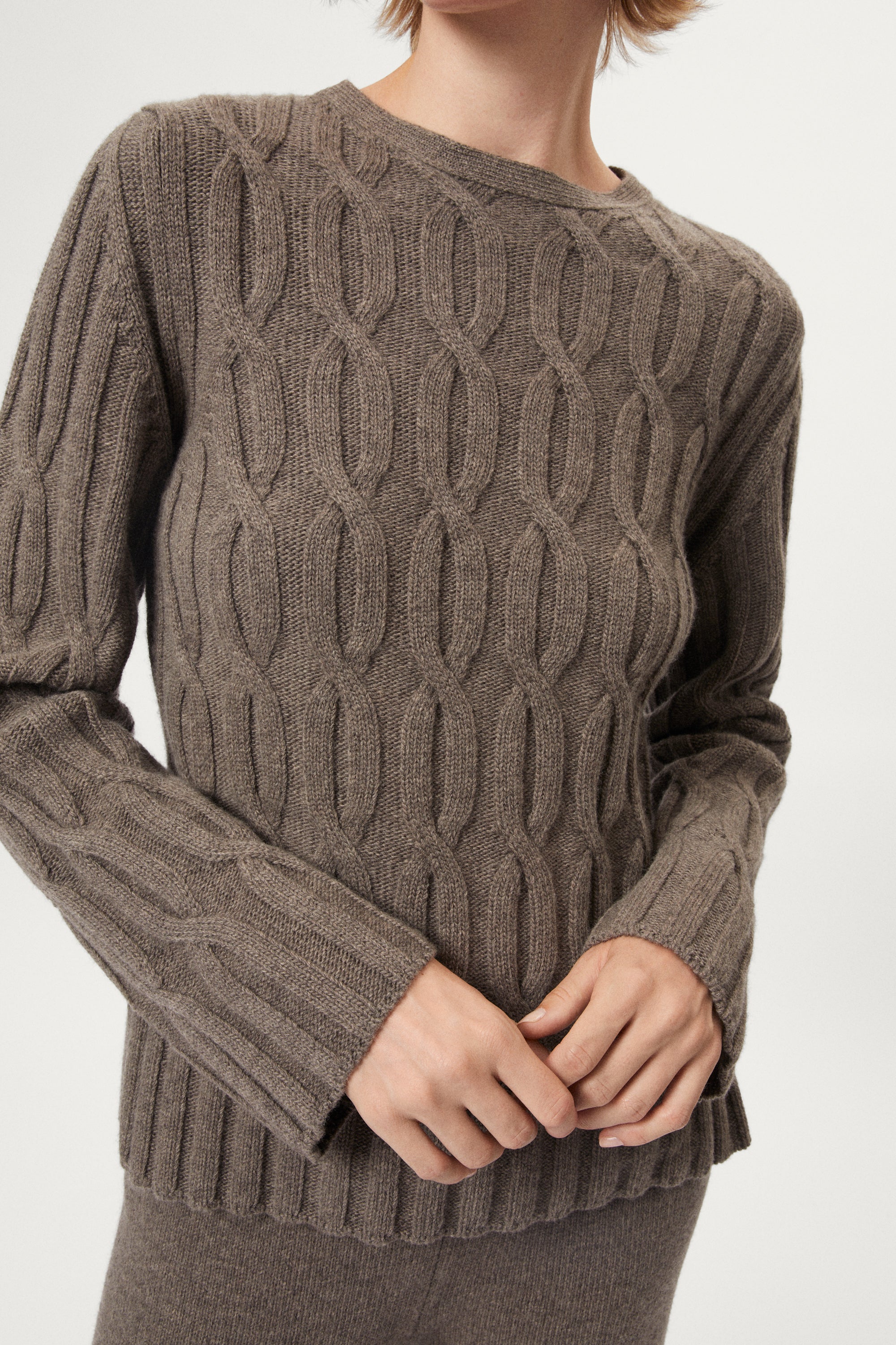 Woman wearing a woolen taupe cable knit sweater and pants on a white background