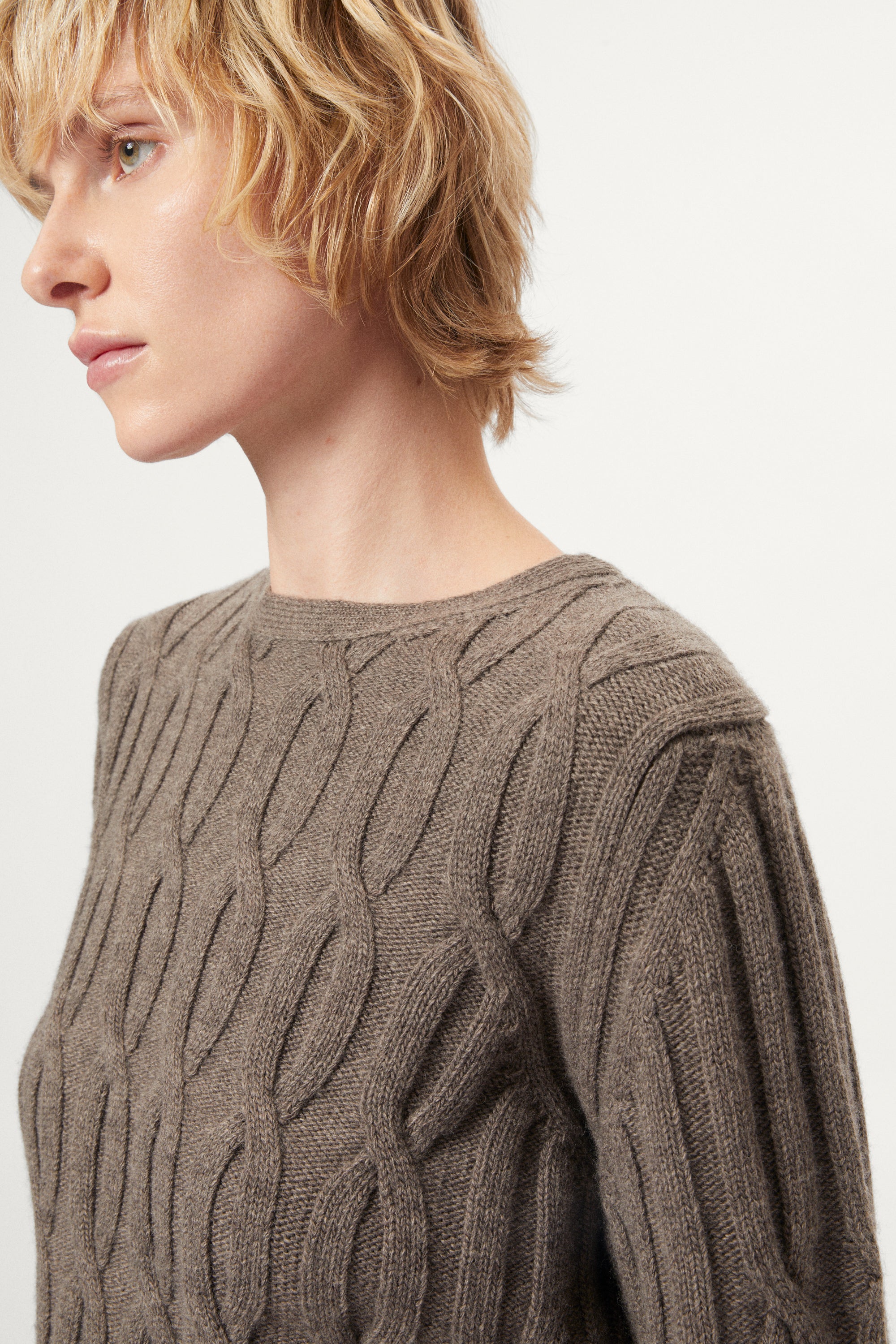 Woman wearing a woolen taupe cable knit sweater and pants on a white background
