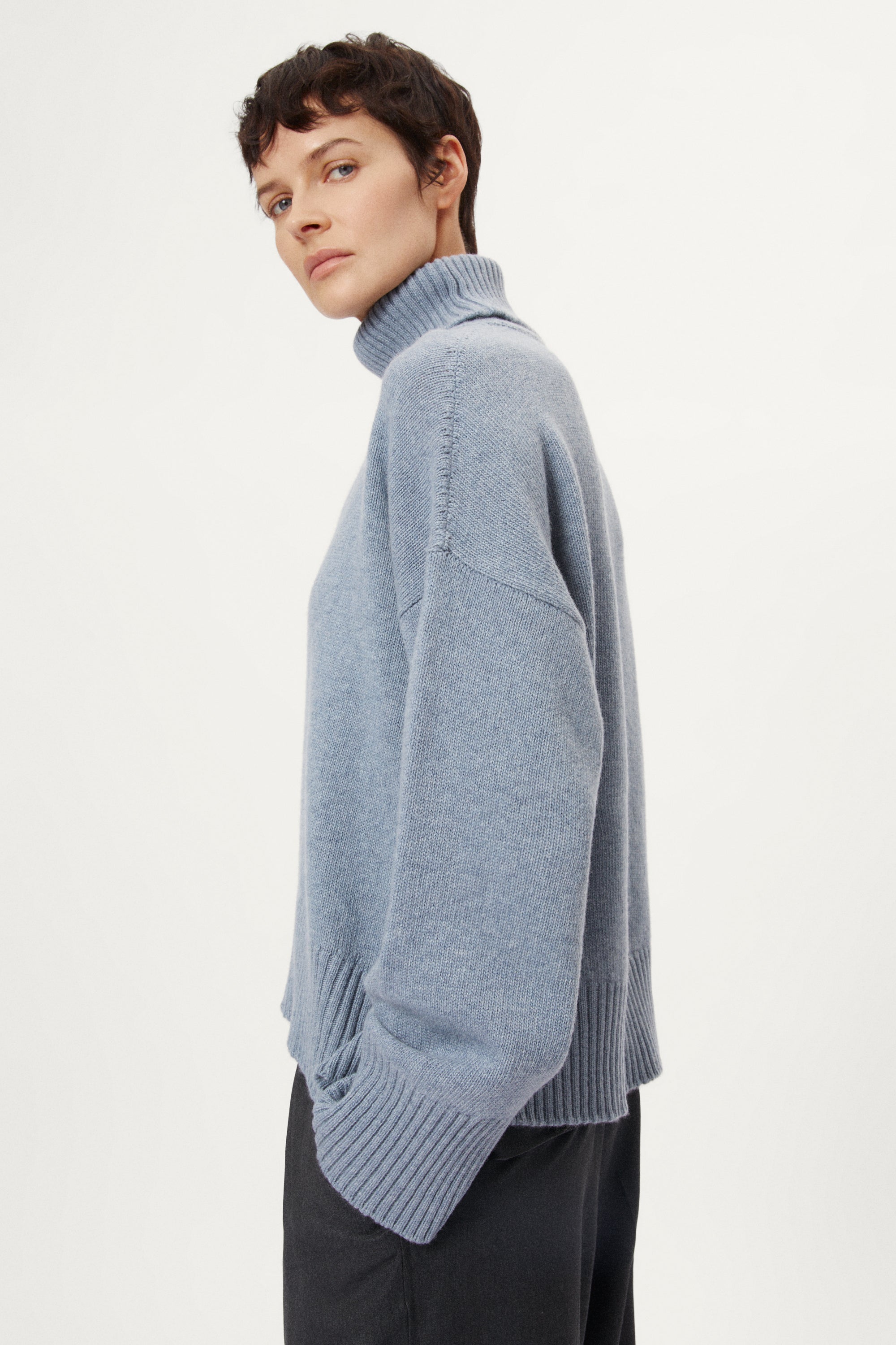 Person wearing a woolen light blue turtleneck sweater and dark pants on a white background