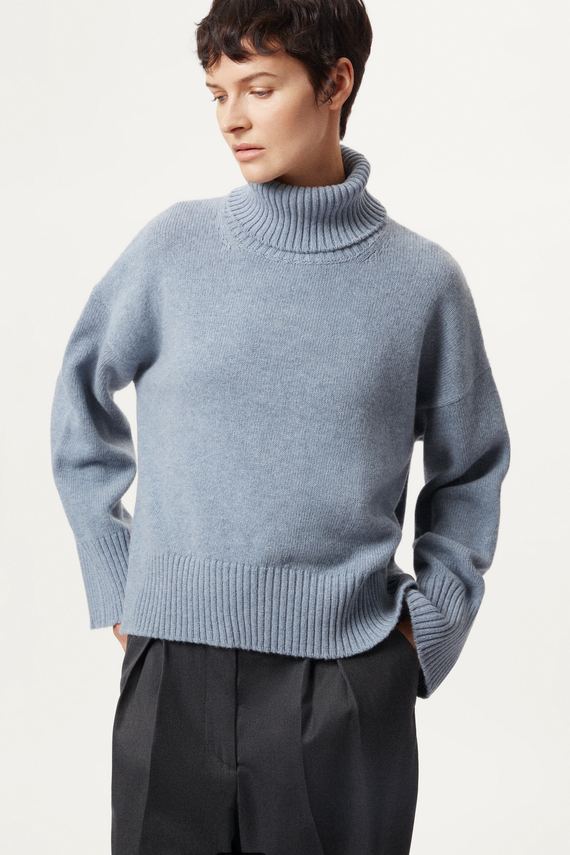 Person wearing a woolen light blue turtleneck sweater and dark pants on a white background