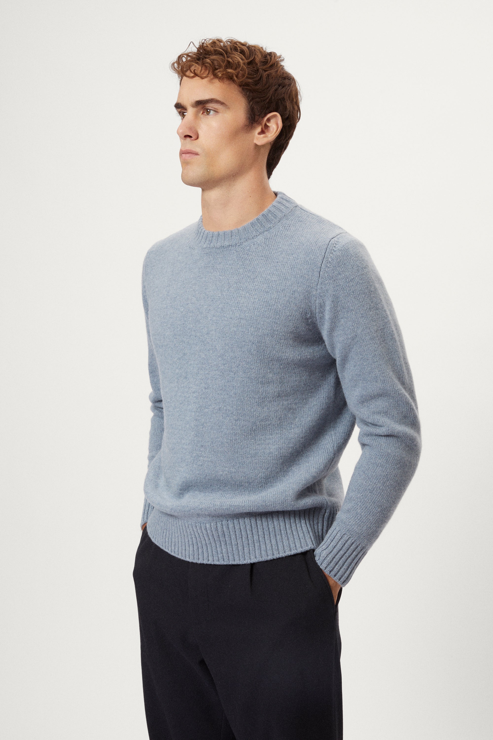 Man wearing a light blue woolen sweater and dark pants on a white background