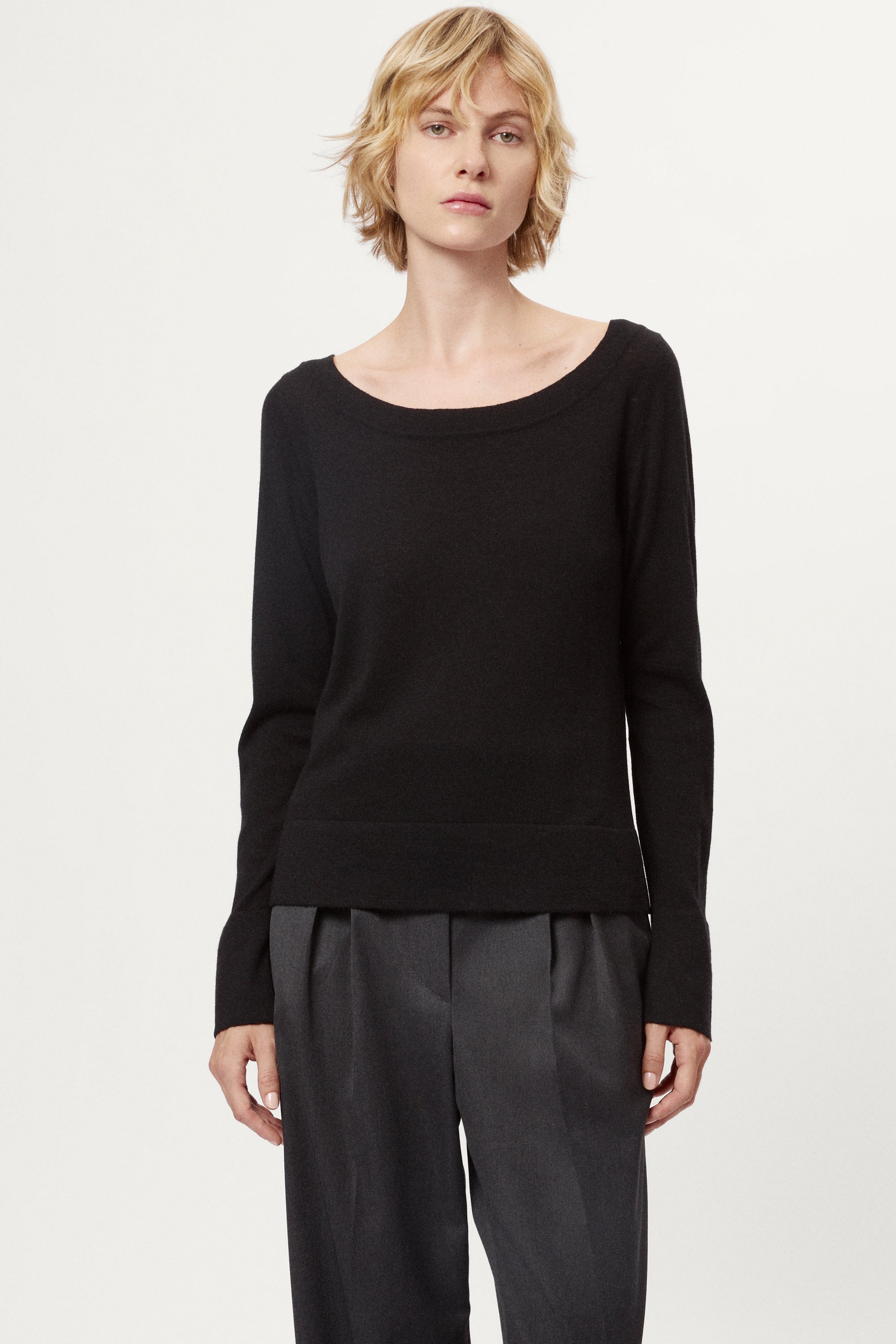 Person wearing a black long-sleeve top and dark gray pleated pants on a white background