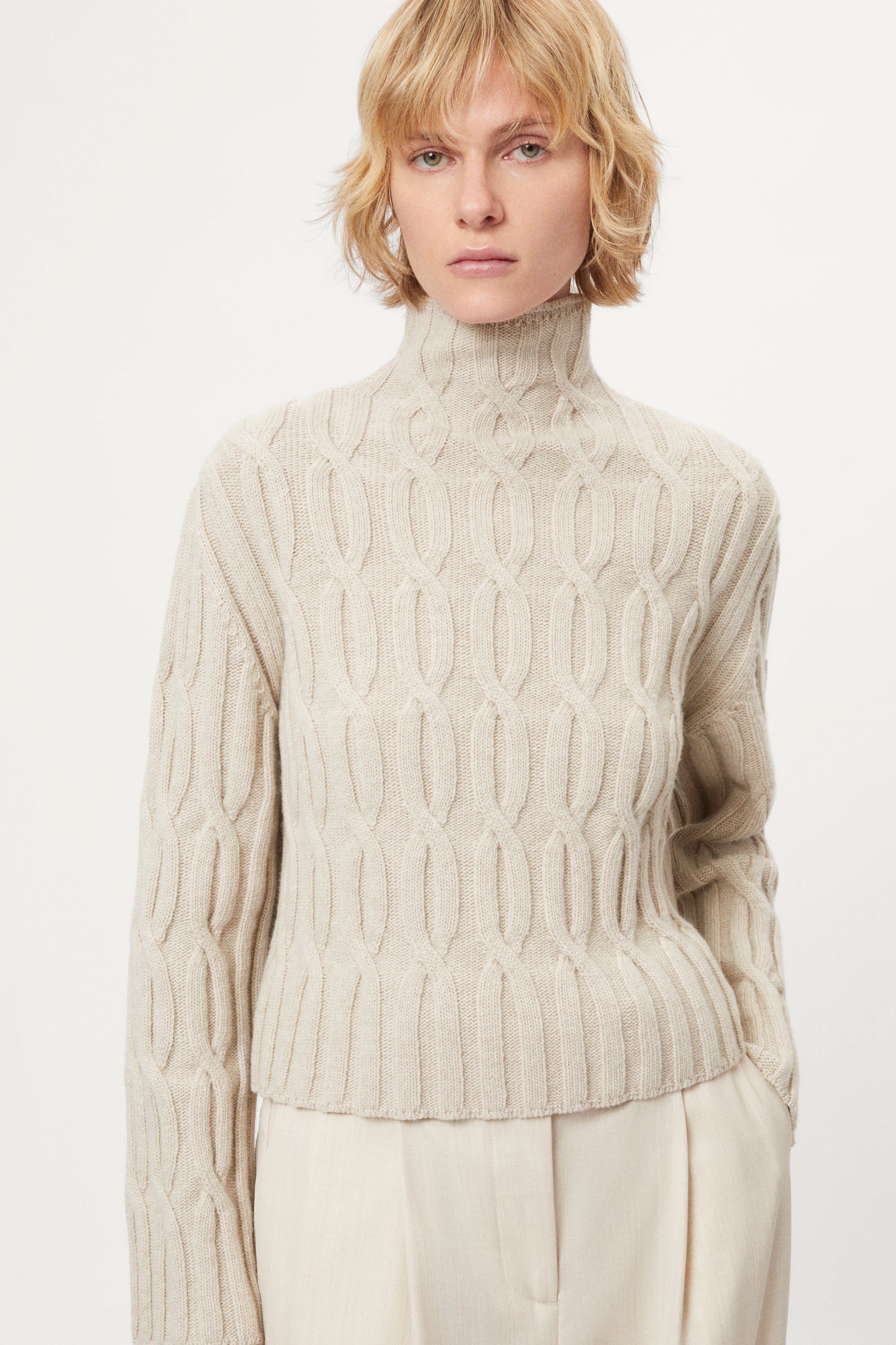 woman wearing a woolen beige cable knit turtleneck sweater on a white background
