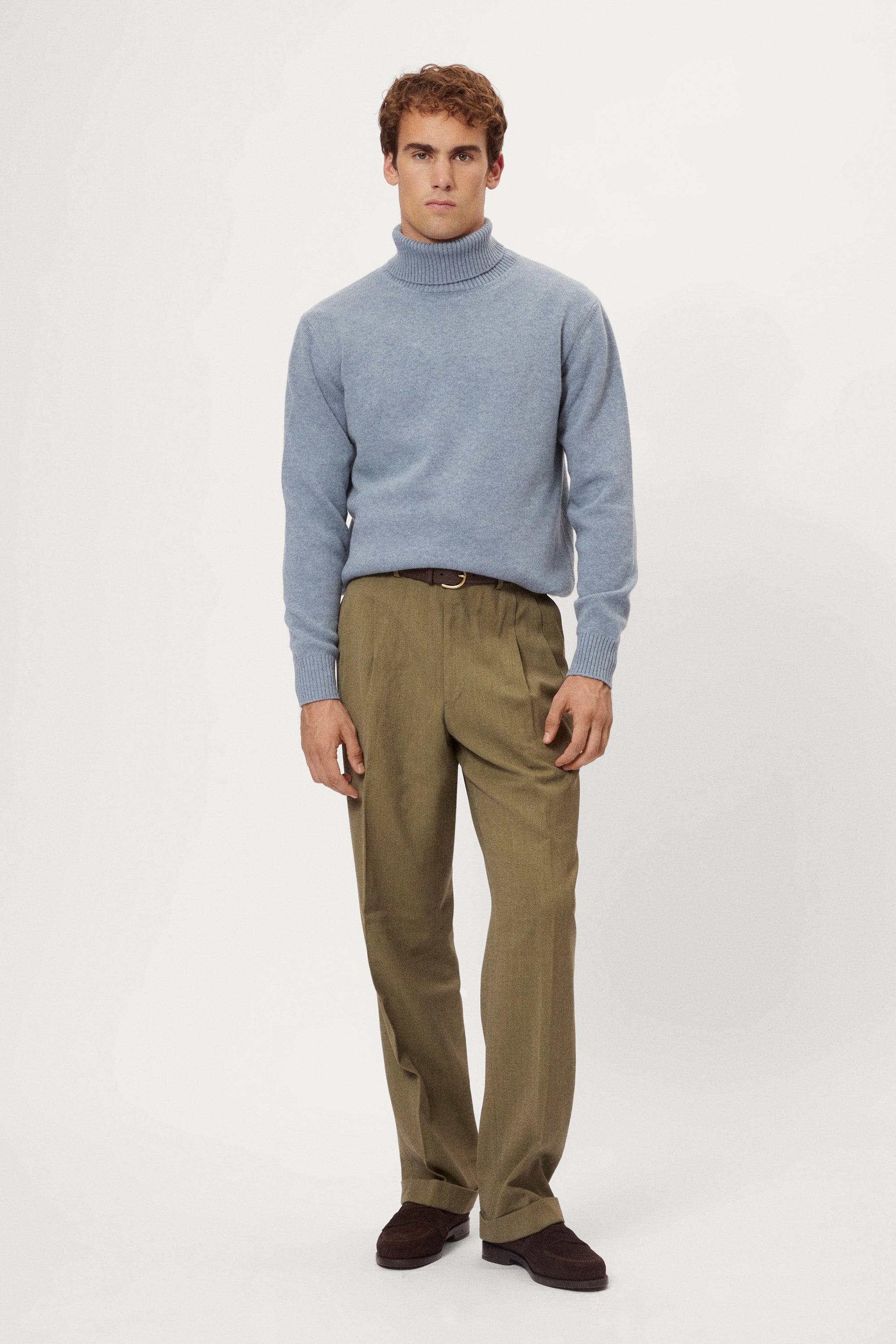 Man wearing a woolen light blue turtleneck sweater and brown pants on a white background
