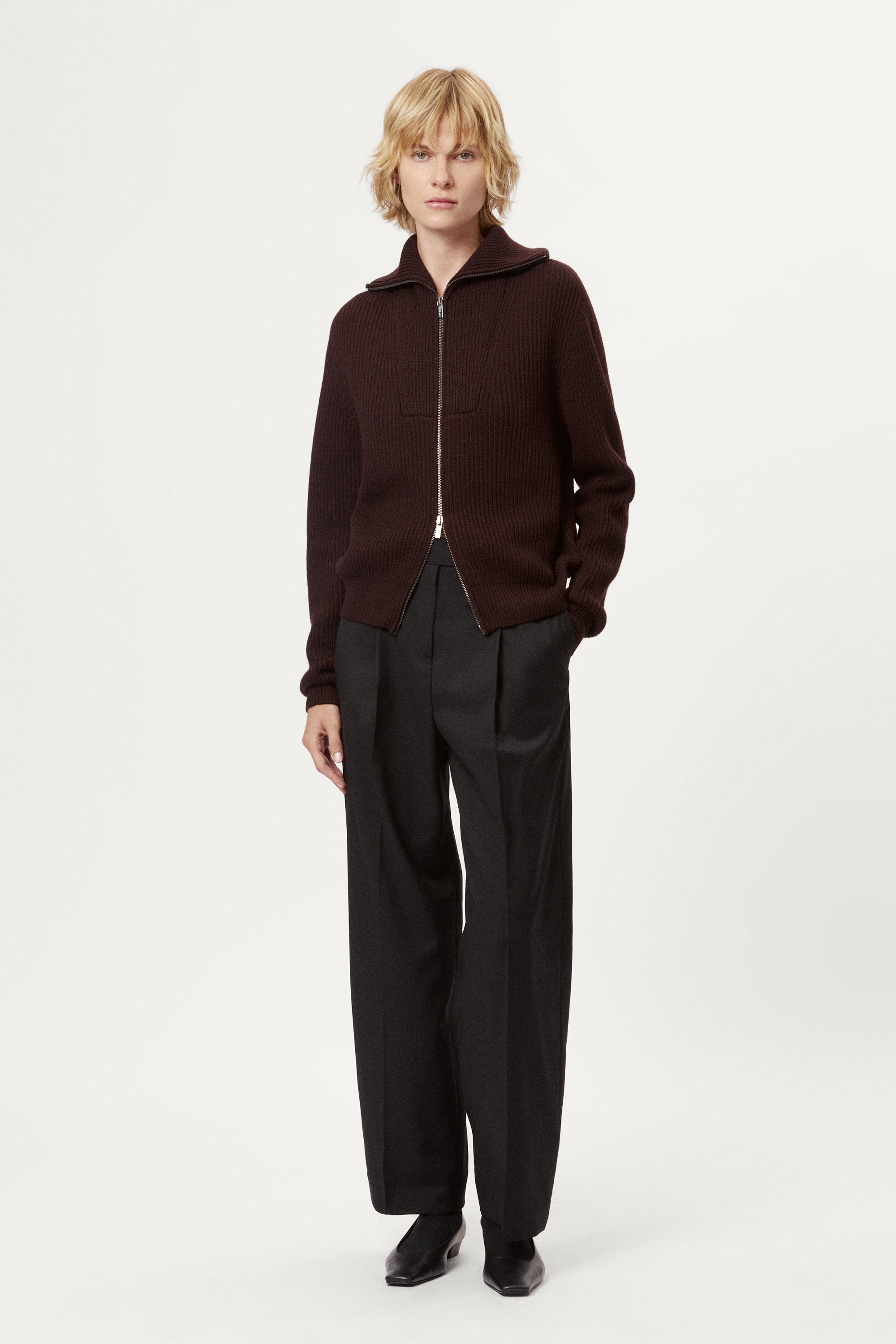 woman wearing a dark brown zip-up sweater and grey pants on a white background