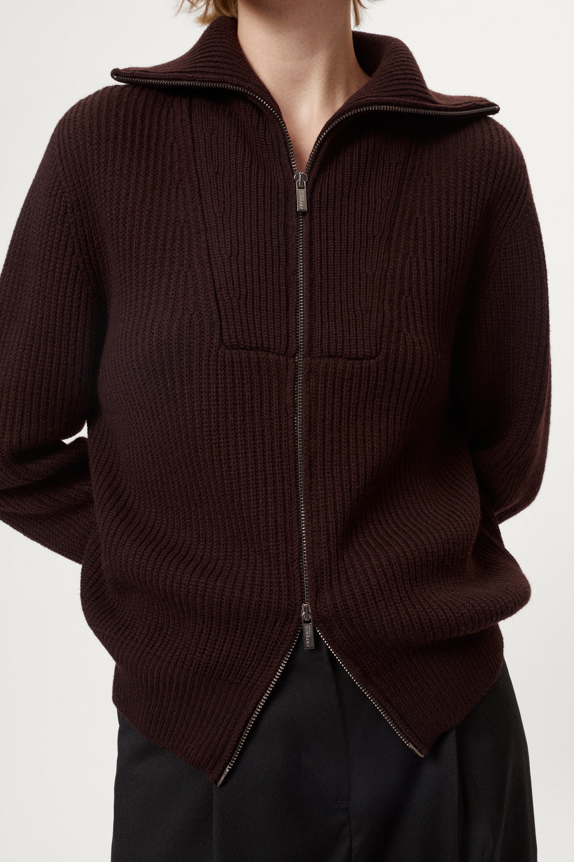 woman wearing a dark brown zip-up sweater and grey pants on a white background