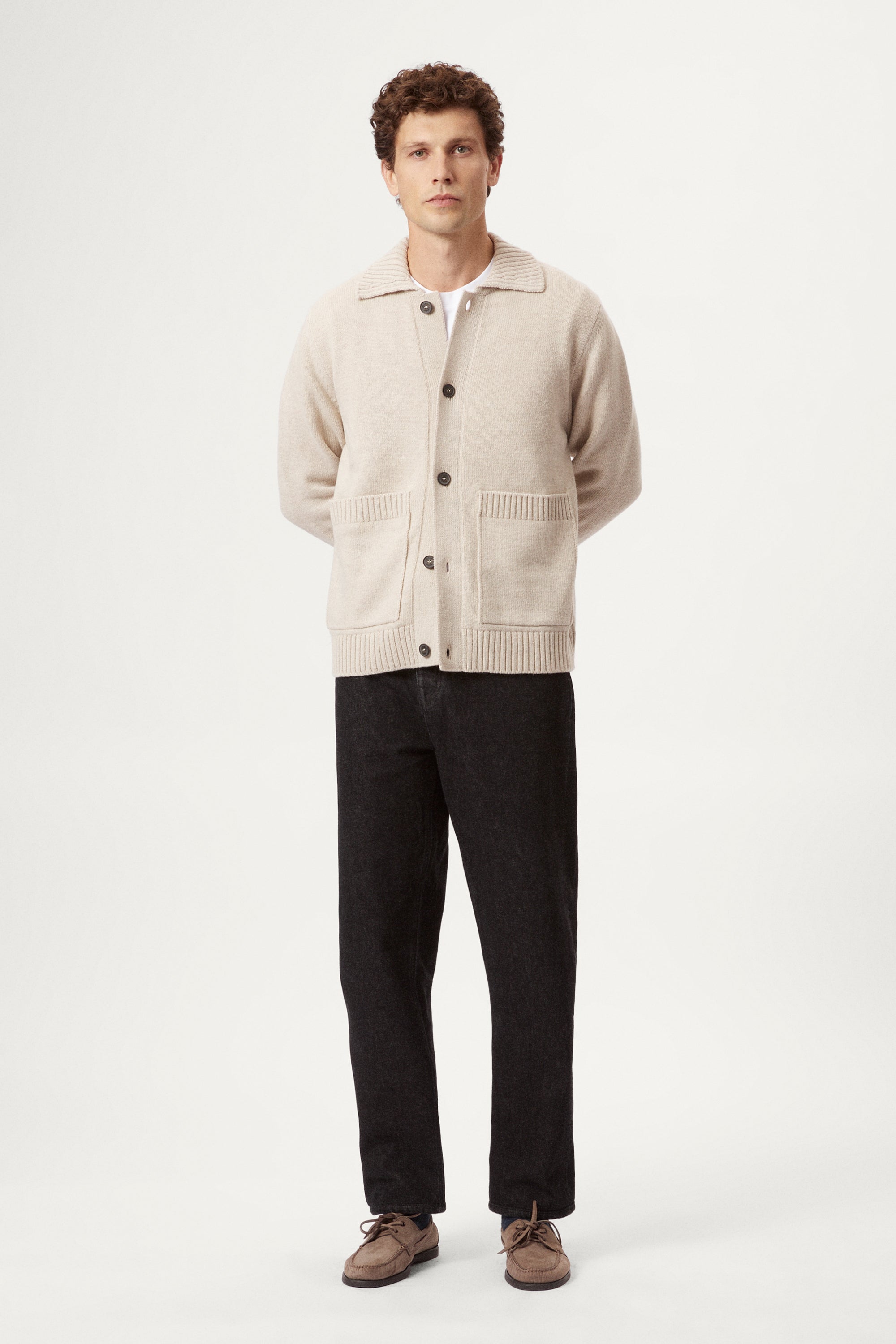 Man wearing a beige woolen cardigan, black pants, and brown shoes on a white background