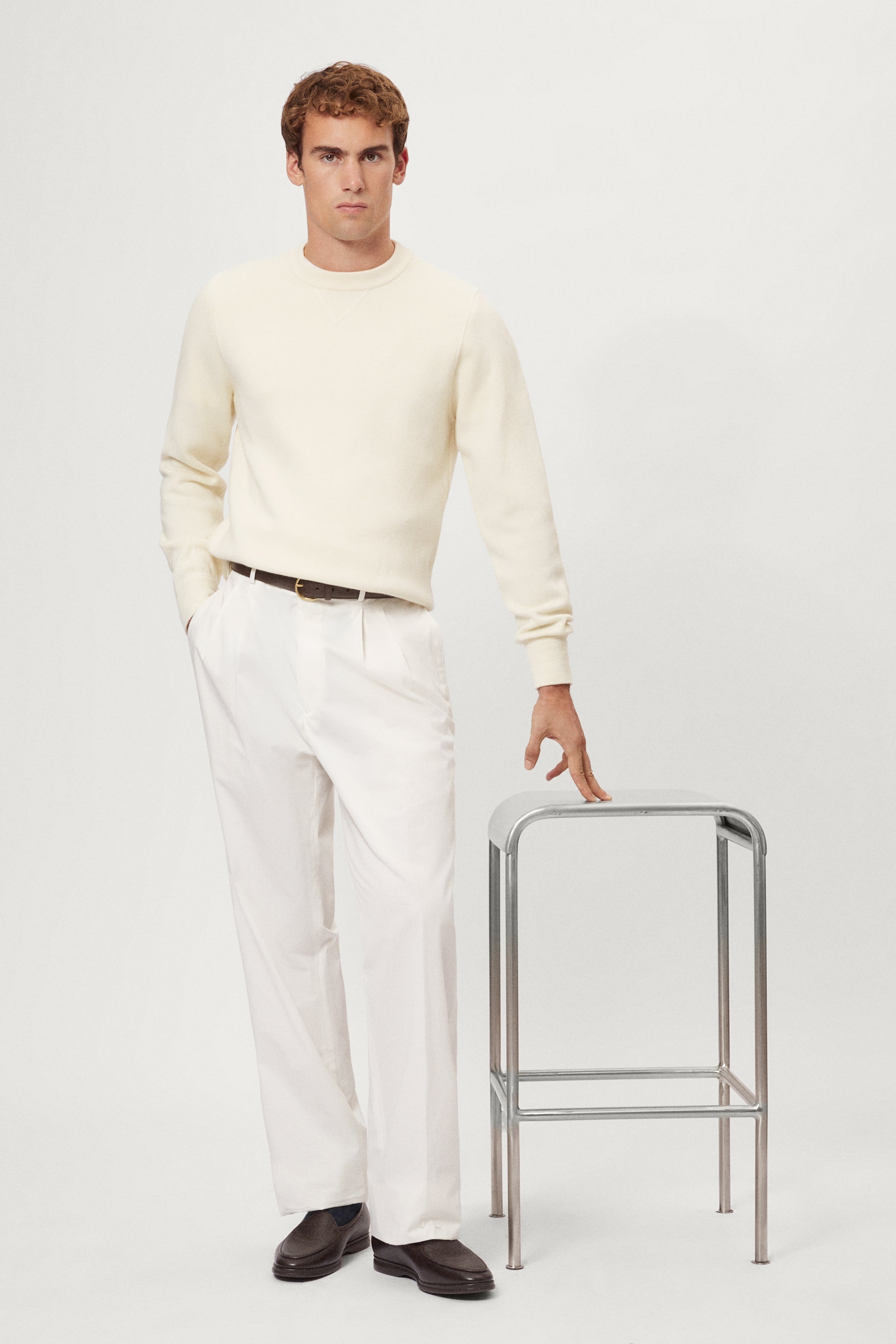 Man in a wool white sweater and white pants holding a metal stool on a plain background