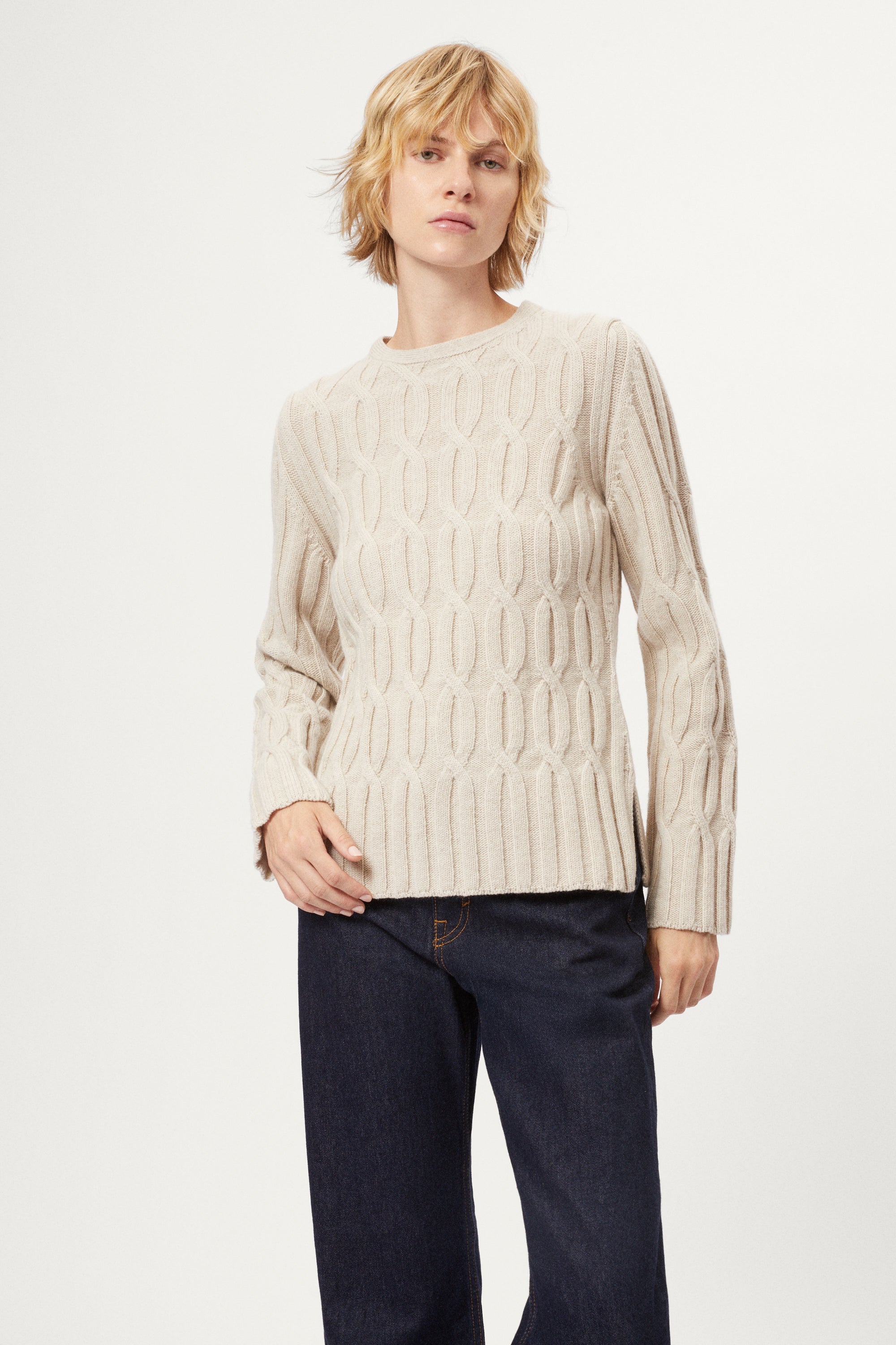Woman wearing an ecru woolen cable knit sweater and dark jeans on a white background
