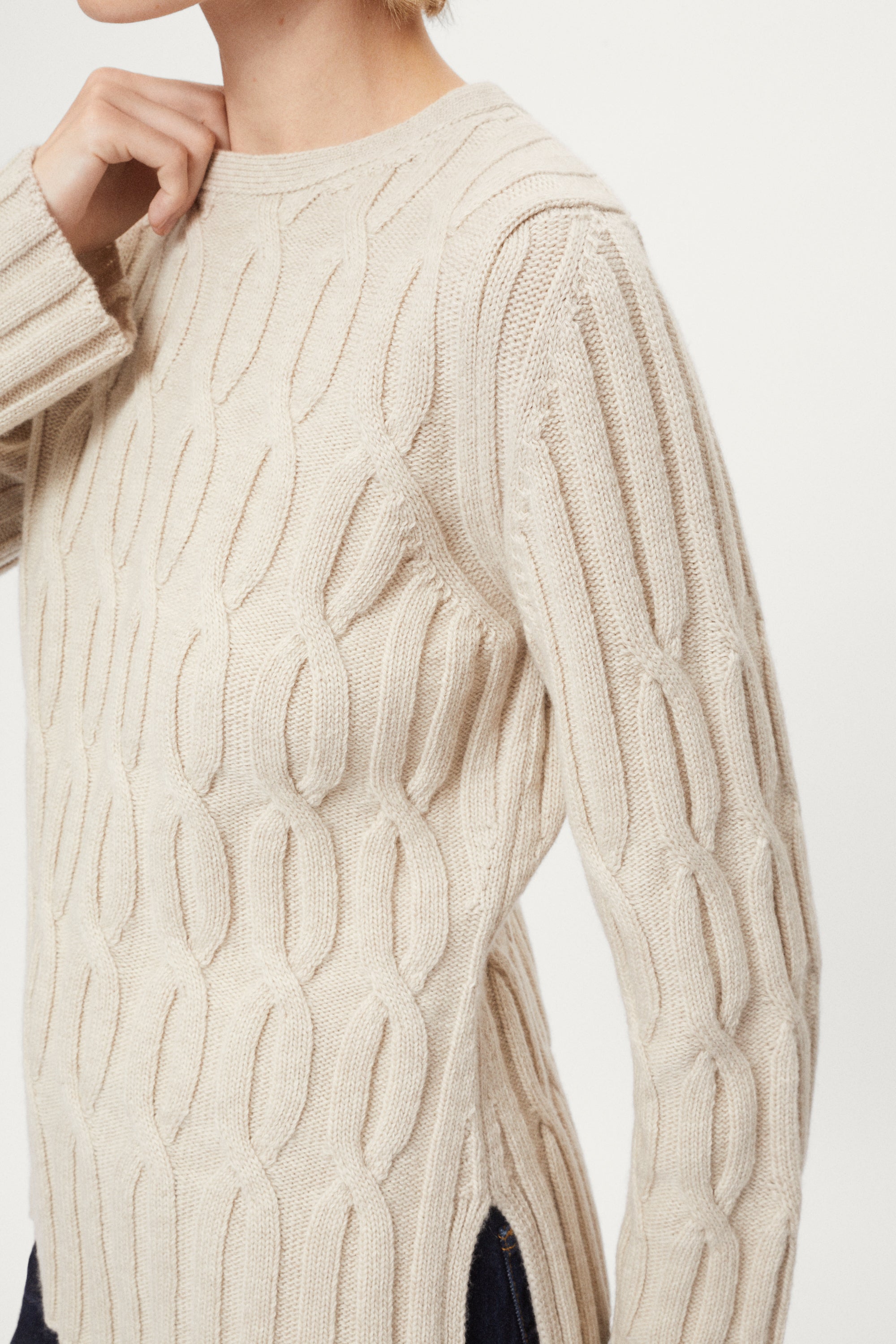 Woman wearing an ecru woolen cable knit sweater and dark jeans on a white background