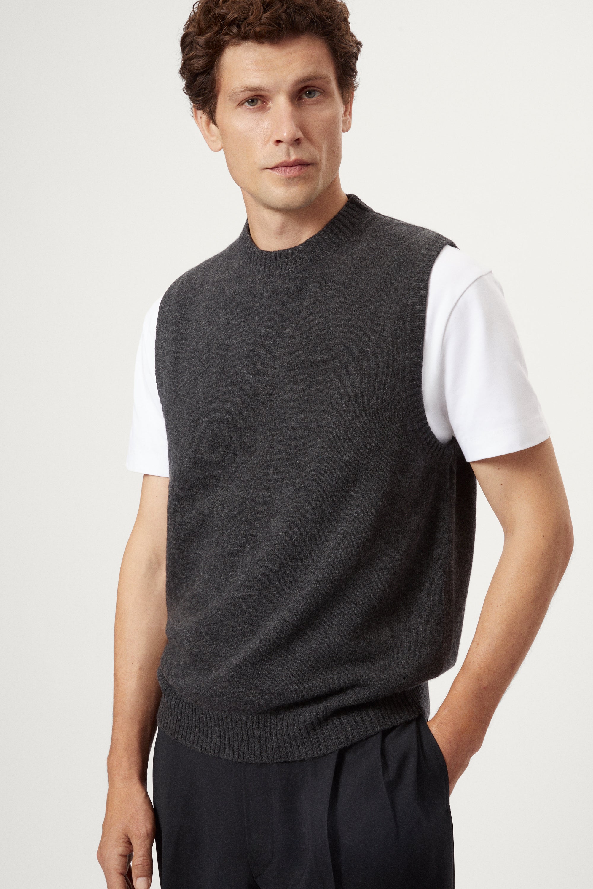 Man wearing a dark gray knitted vest over a white shirt on a light background