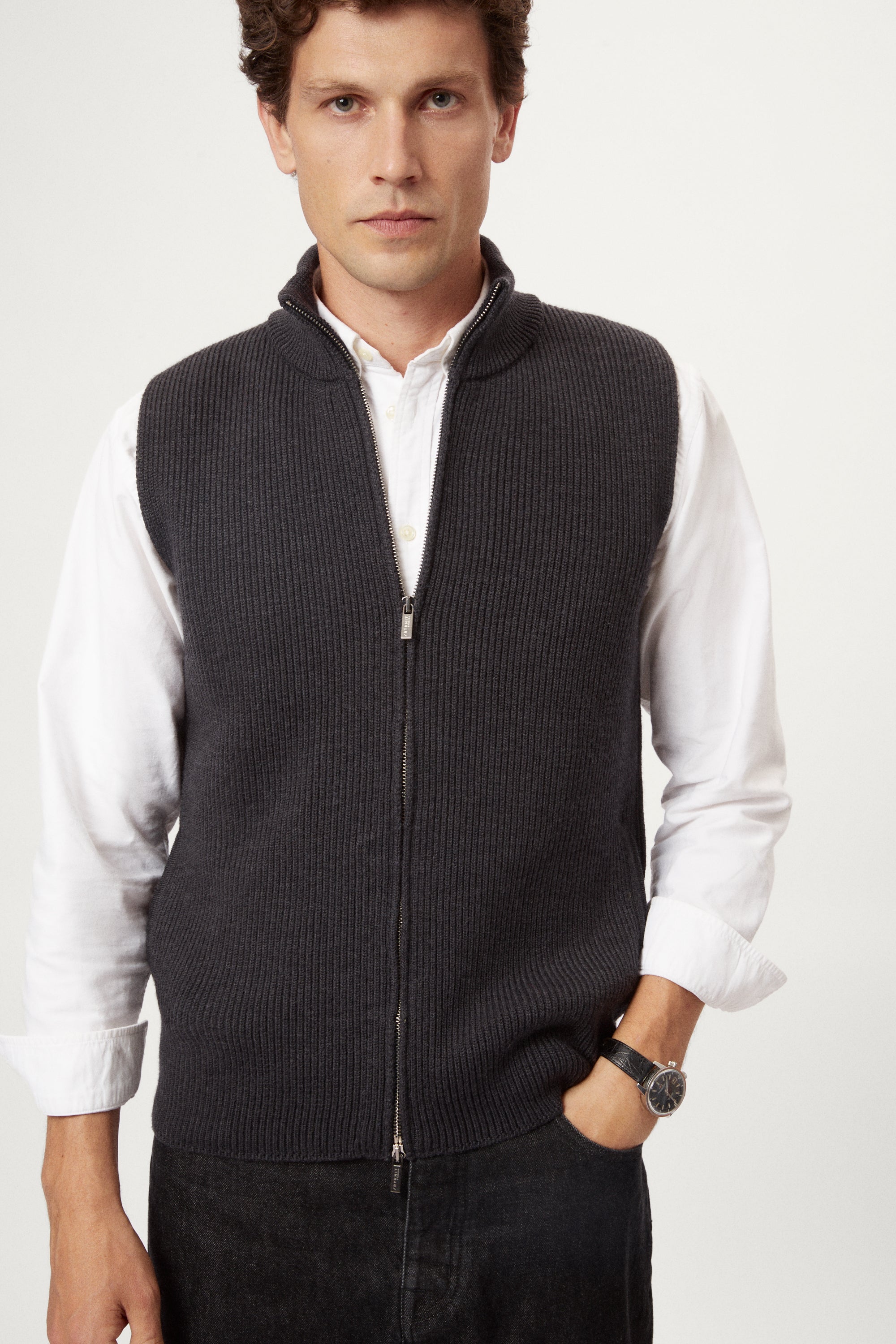 Man wearing a dark gray zip-up vest over a white shirt on a white background