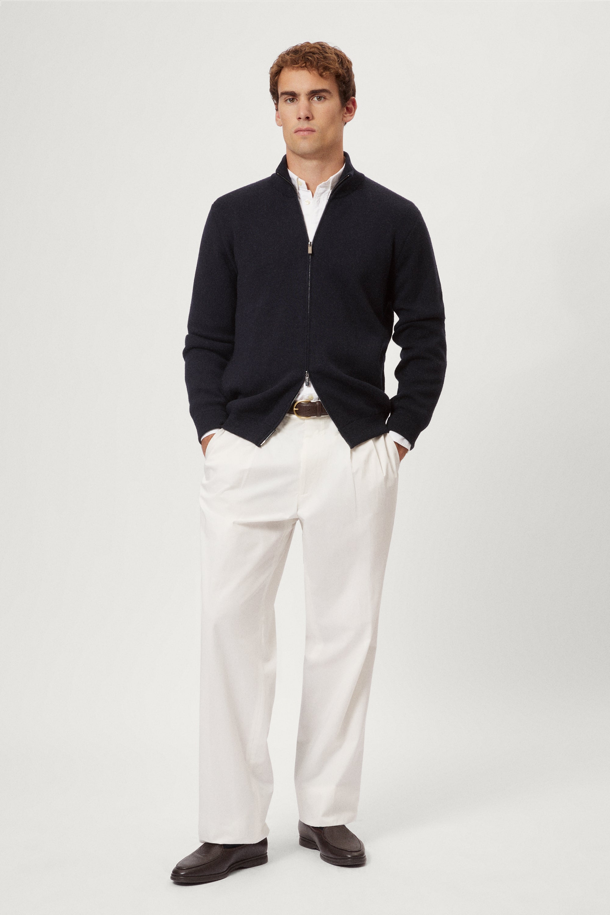 Man wearing a navy blue zip-up sweater over a white shirt with light-colored pants on a plain background