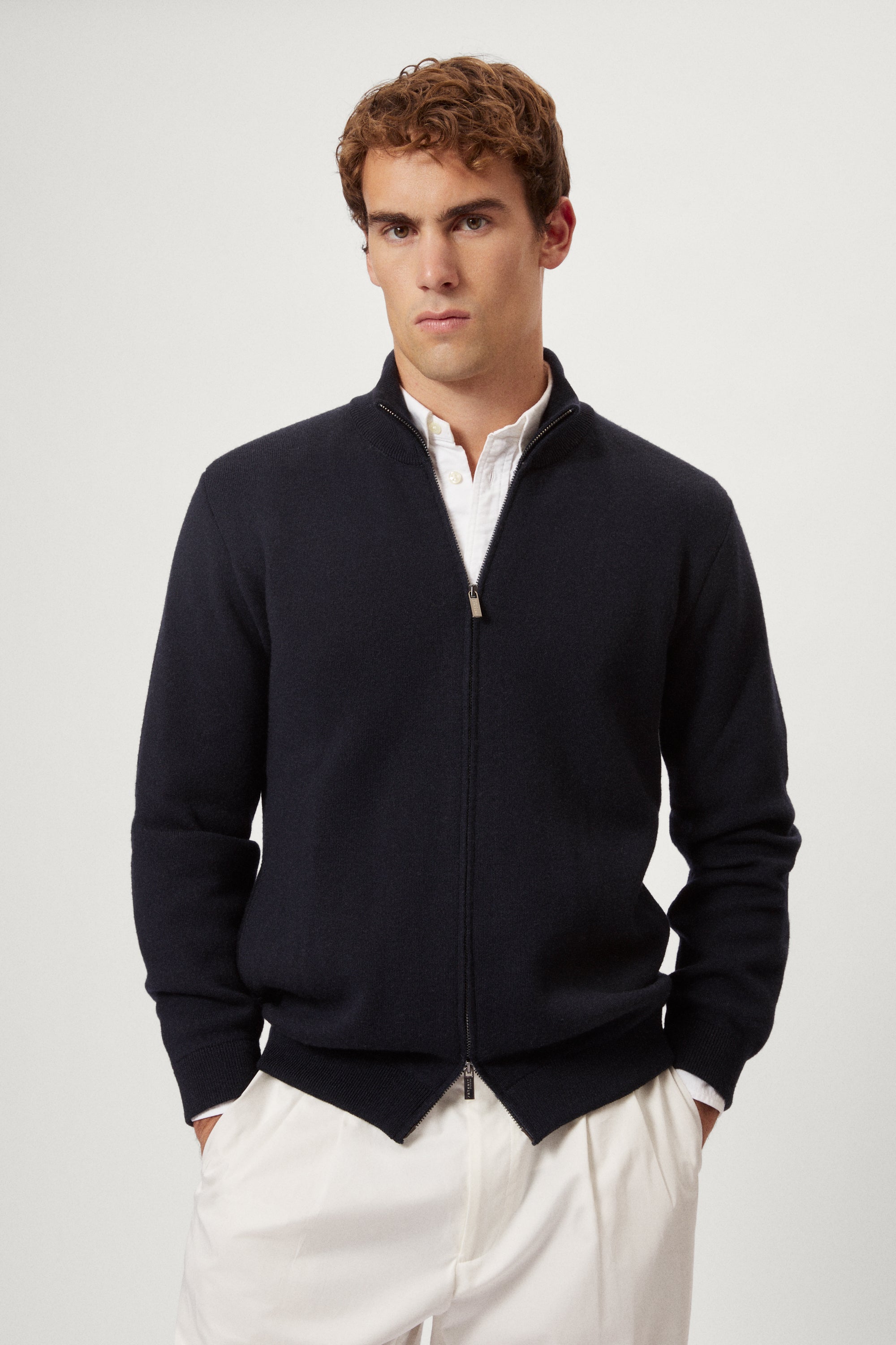 Man wearing a navy blue zip-up sweater over a white shirt with light-colored pants on a plain background