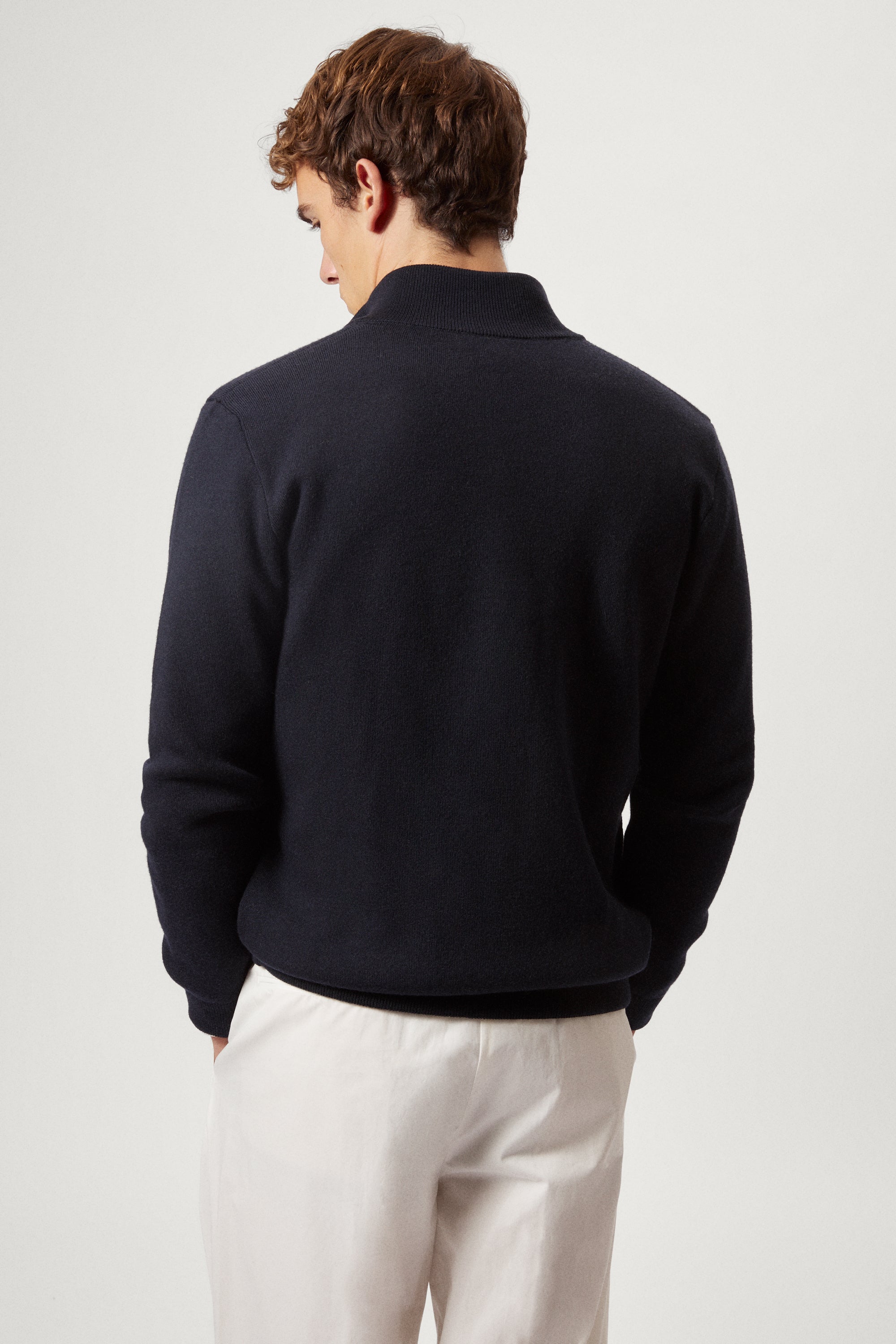 Man wearing a navy blue zip-up sweater over a white shirt with light-colored pants on a plain background