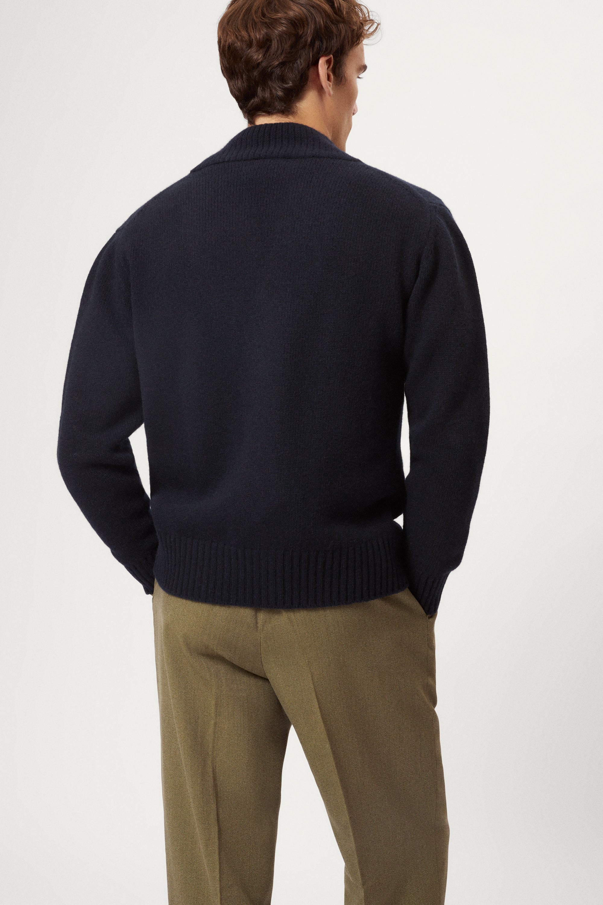 Man wearing a woolen blue cardigan over a white shirt with beige pants on a white background