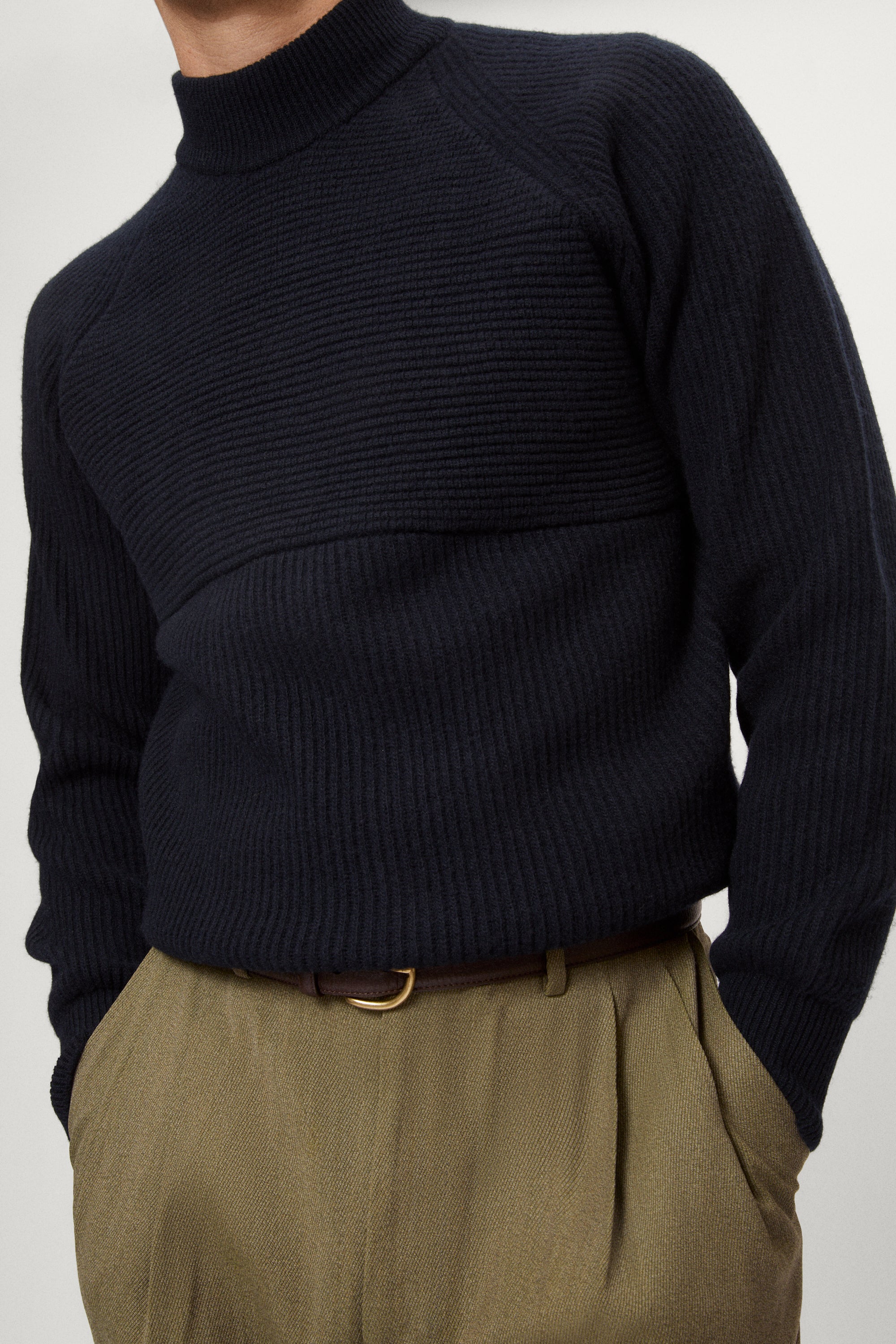 Man wearing a dark blue woolen ribbed sweater and green pants on a white background
