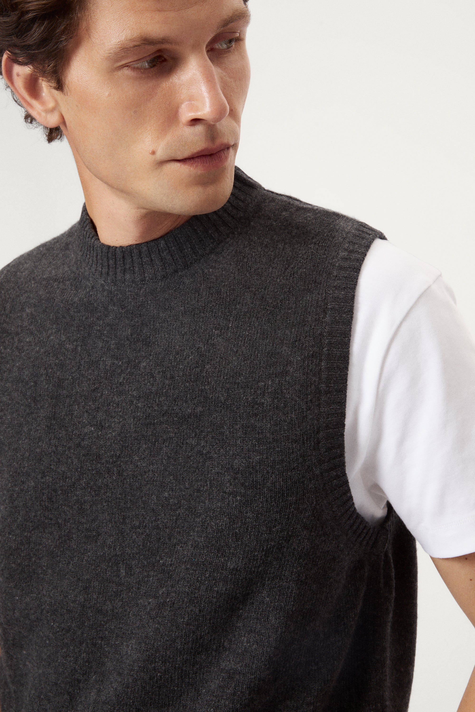 Man wearing a dark gray knitted vest over a white shirt on a light background
