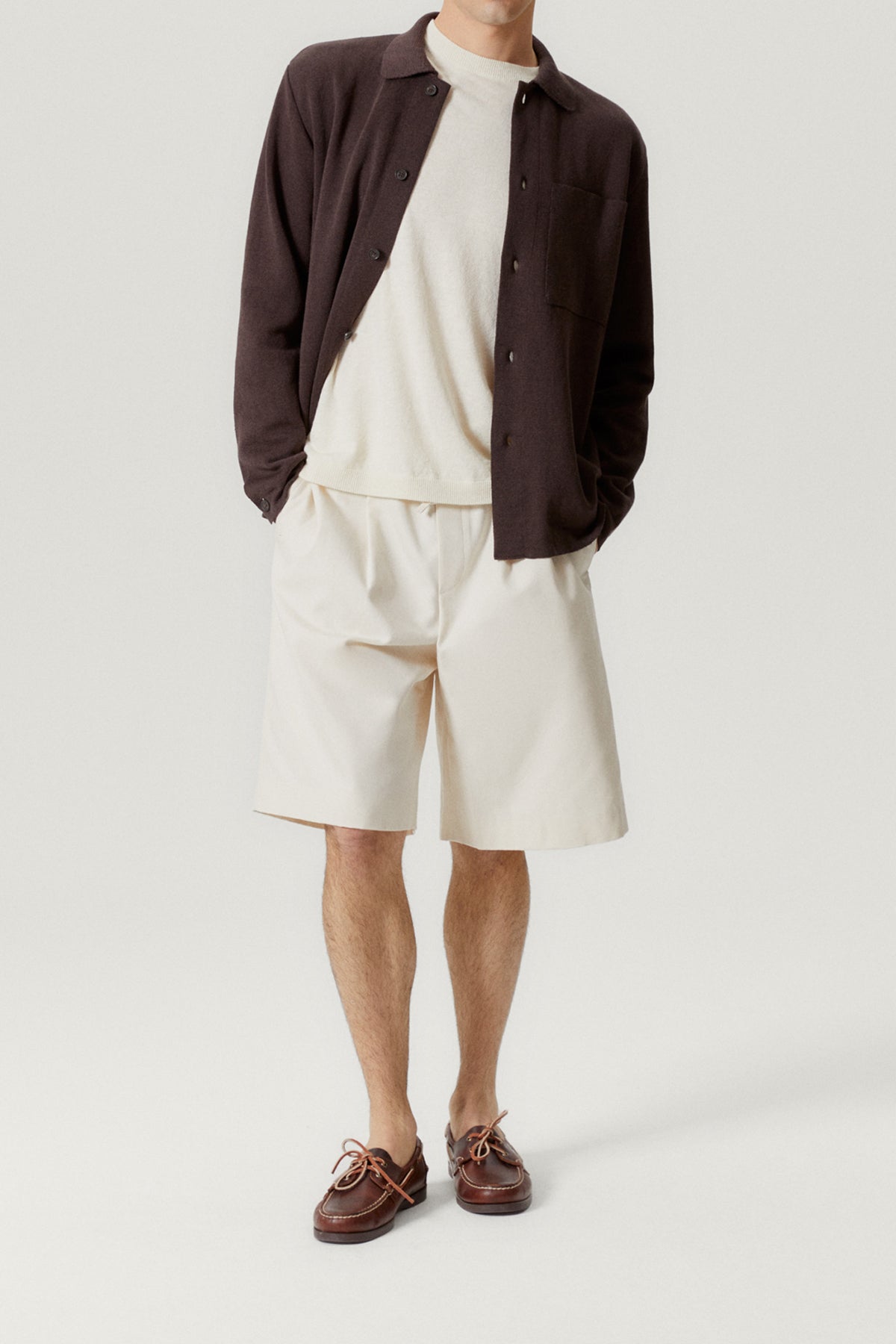 Person wearing a brown overshirt, white sweater, beige shorts, and brown shoes on a light gray background