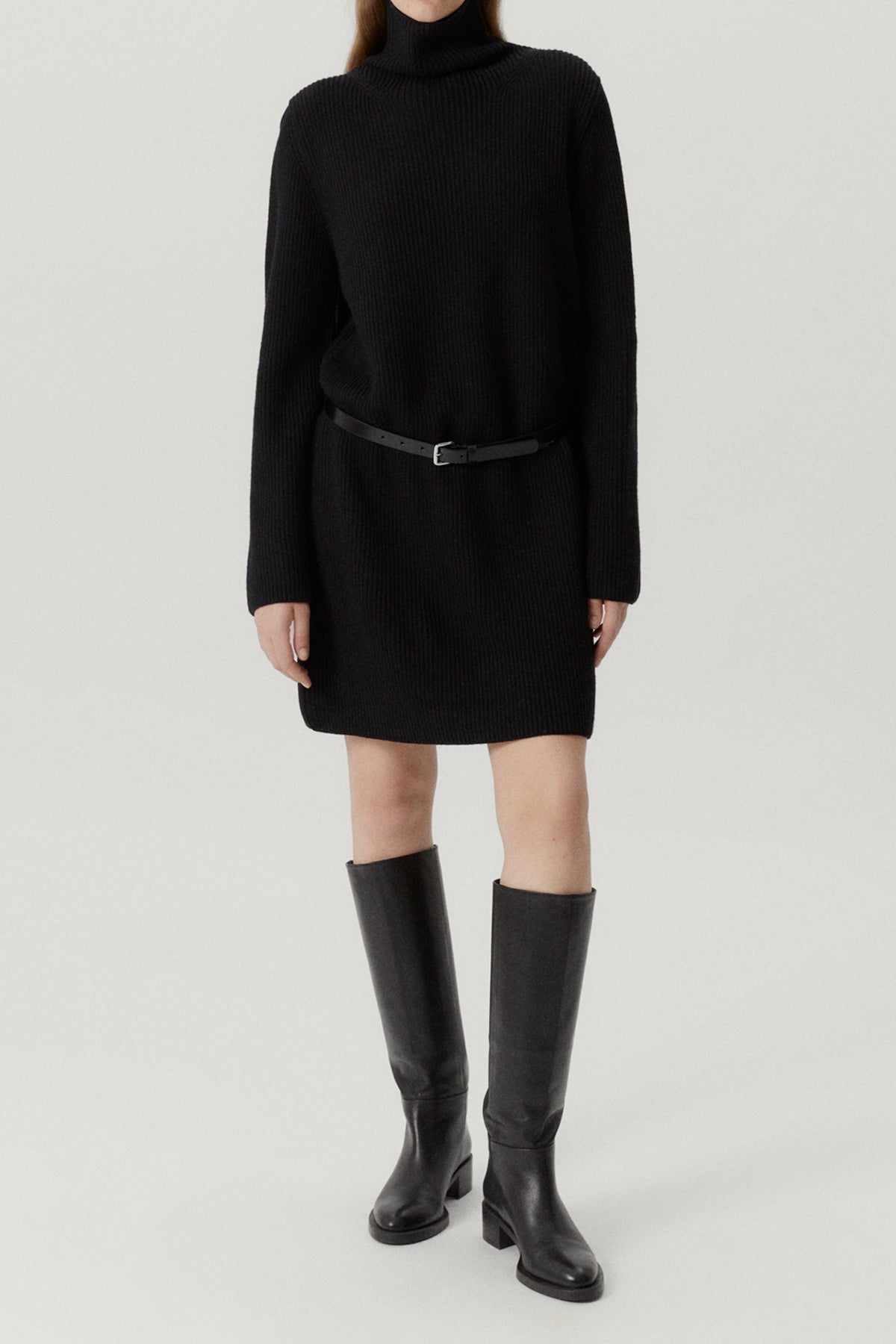 Person wearing a black turtleneck dress and knee-high boots on a light gray background