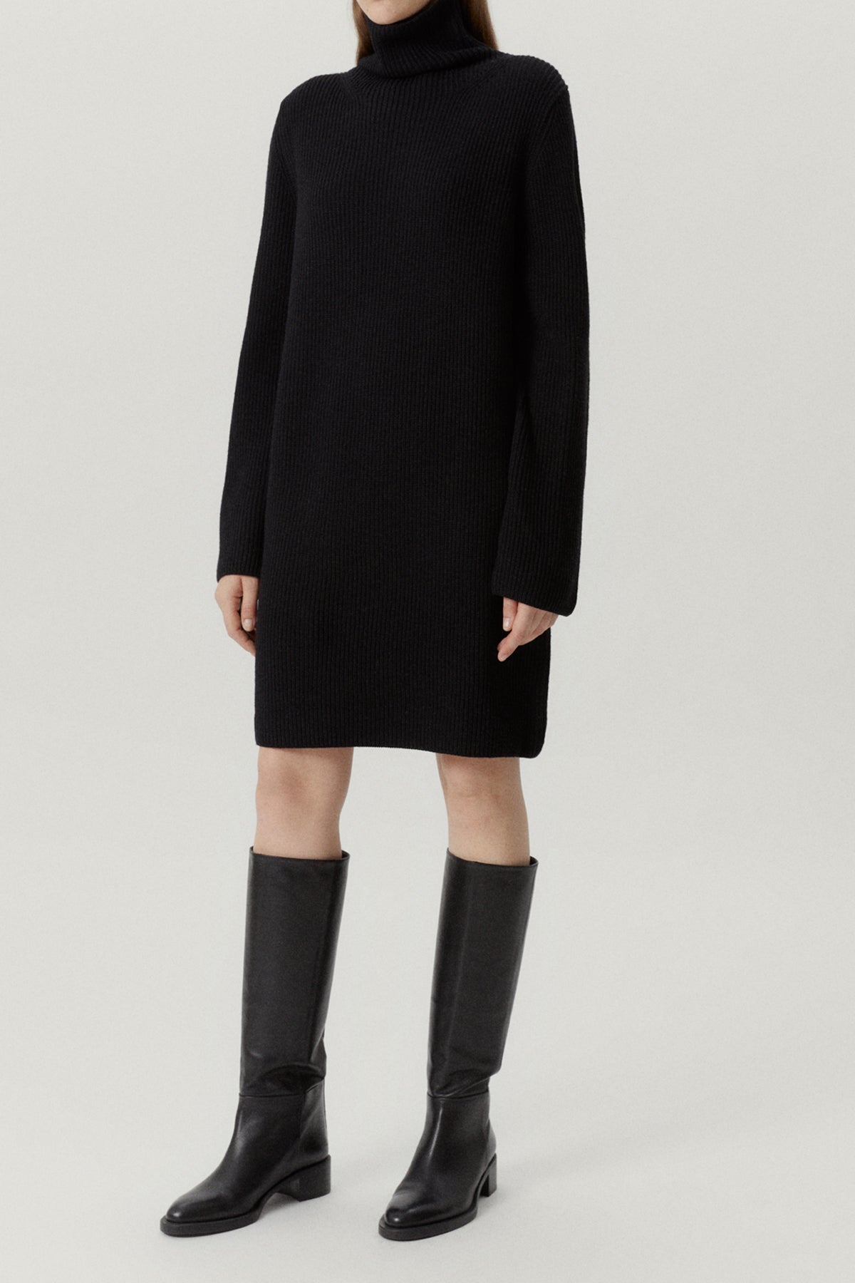Person wearing a black turtleneck dress and knee-high boots on a light gray background