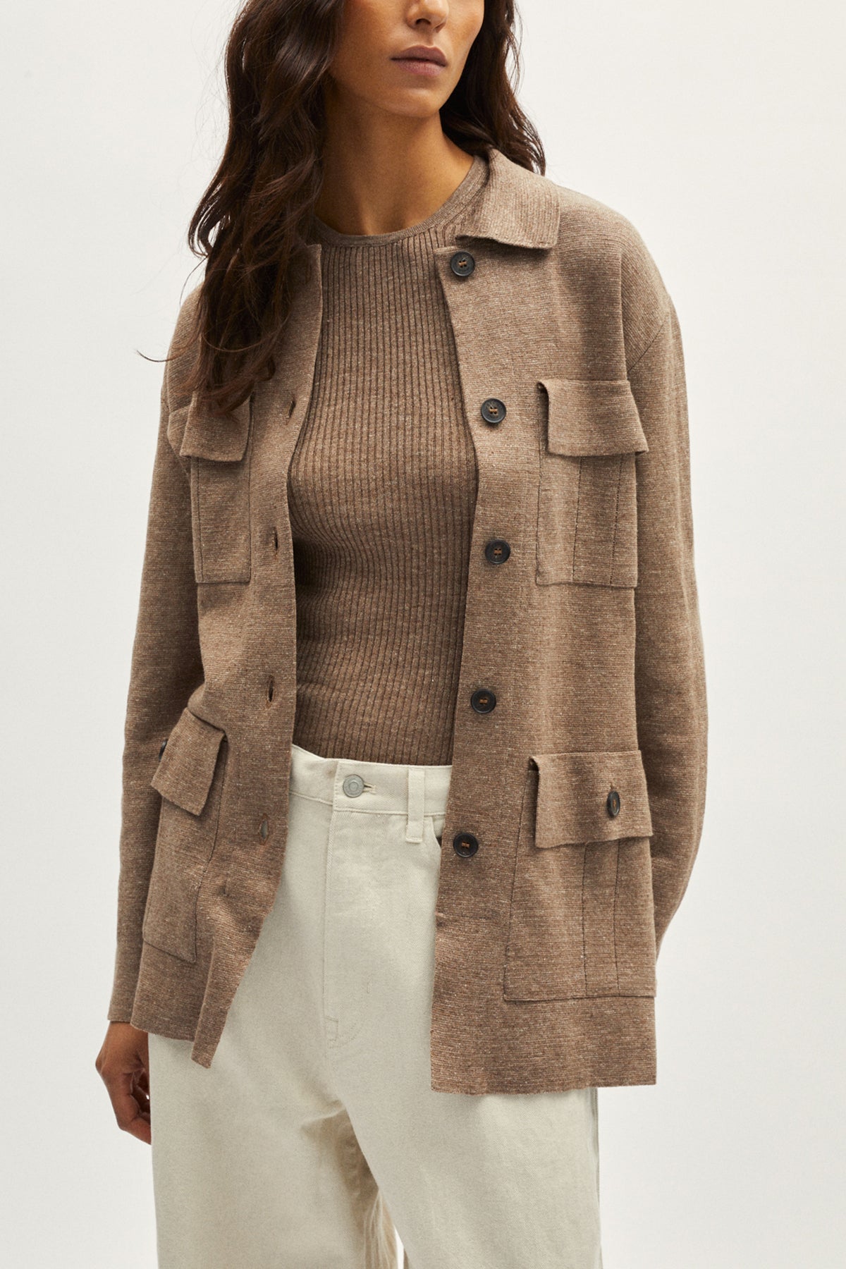 Woman wearing a brown jacket over a brown sweater and white pants on a white background