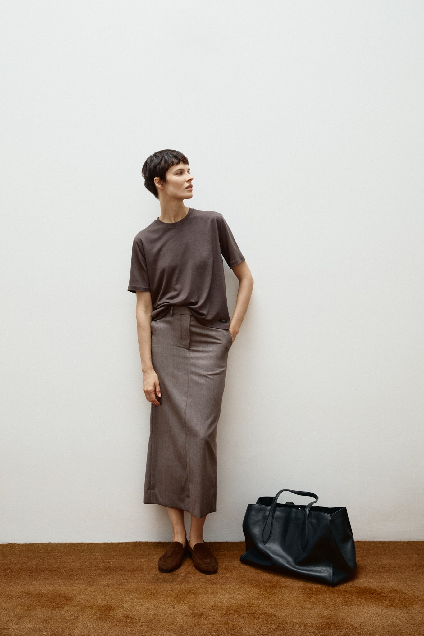 woman wearing a brown wool t-shirt over a taupe skirt standing next to a black bag against a white wall.