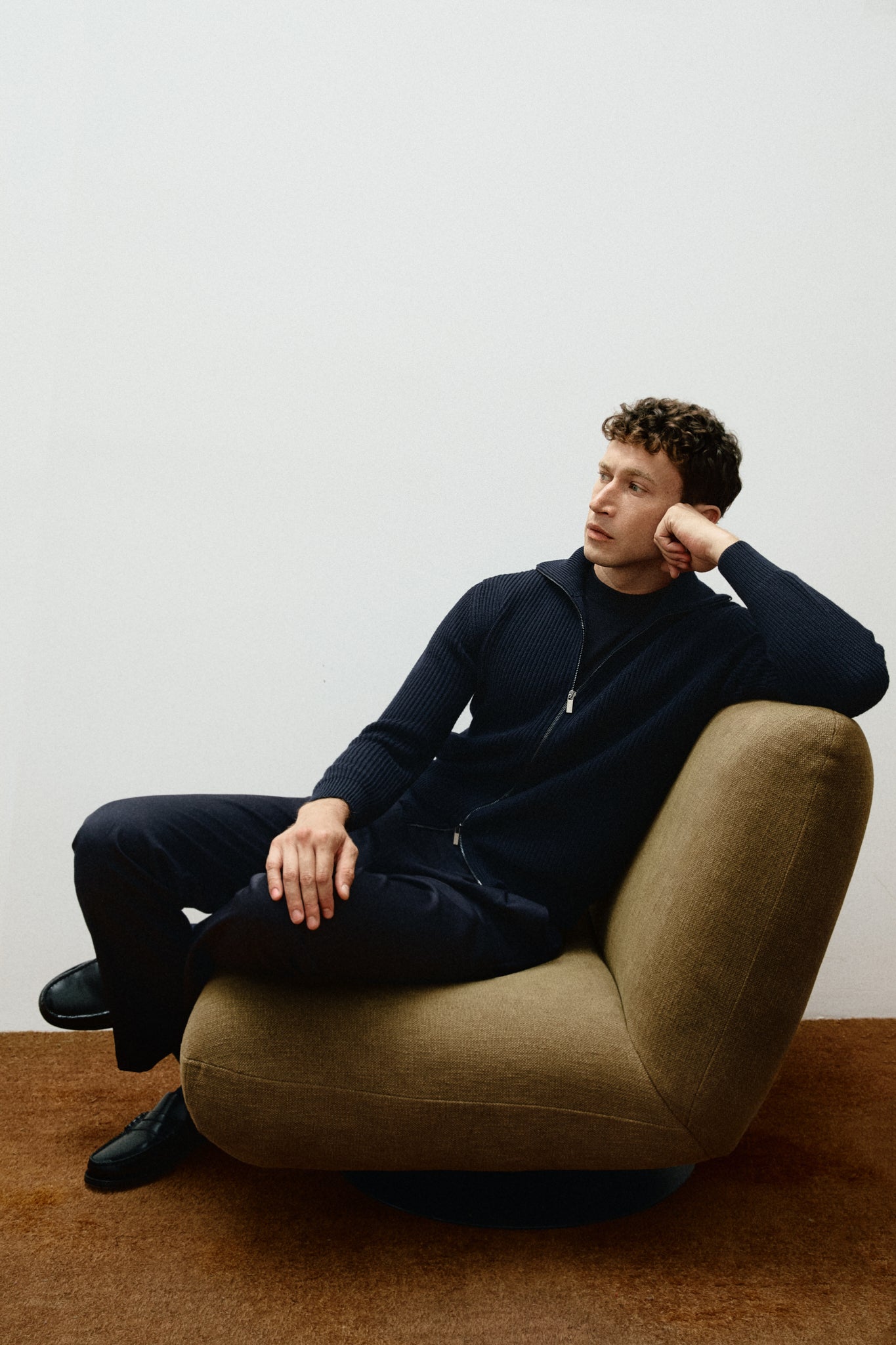 Man wearing a dark blue zip-up sweater  sitting on a beige armchair against a plain white wall