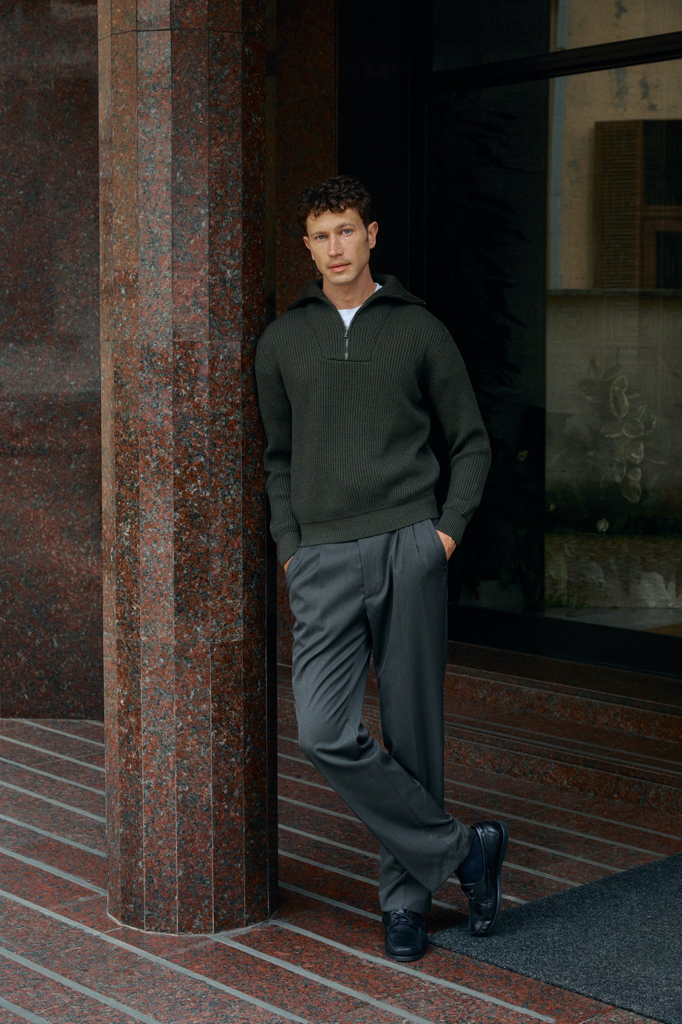 Man in a dark sweater and gray pants leaning against a textured wall.