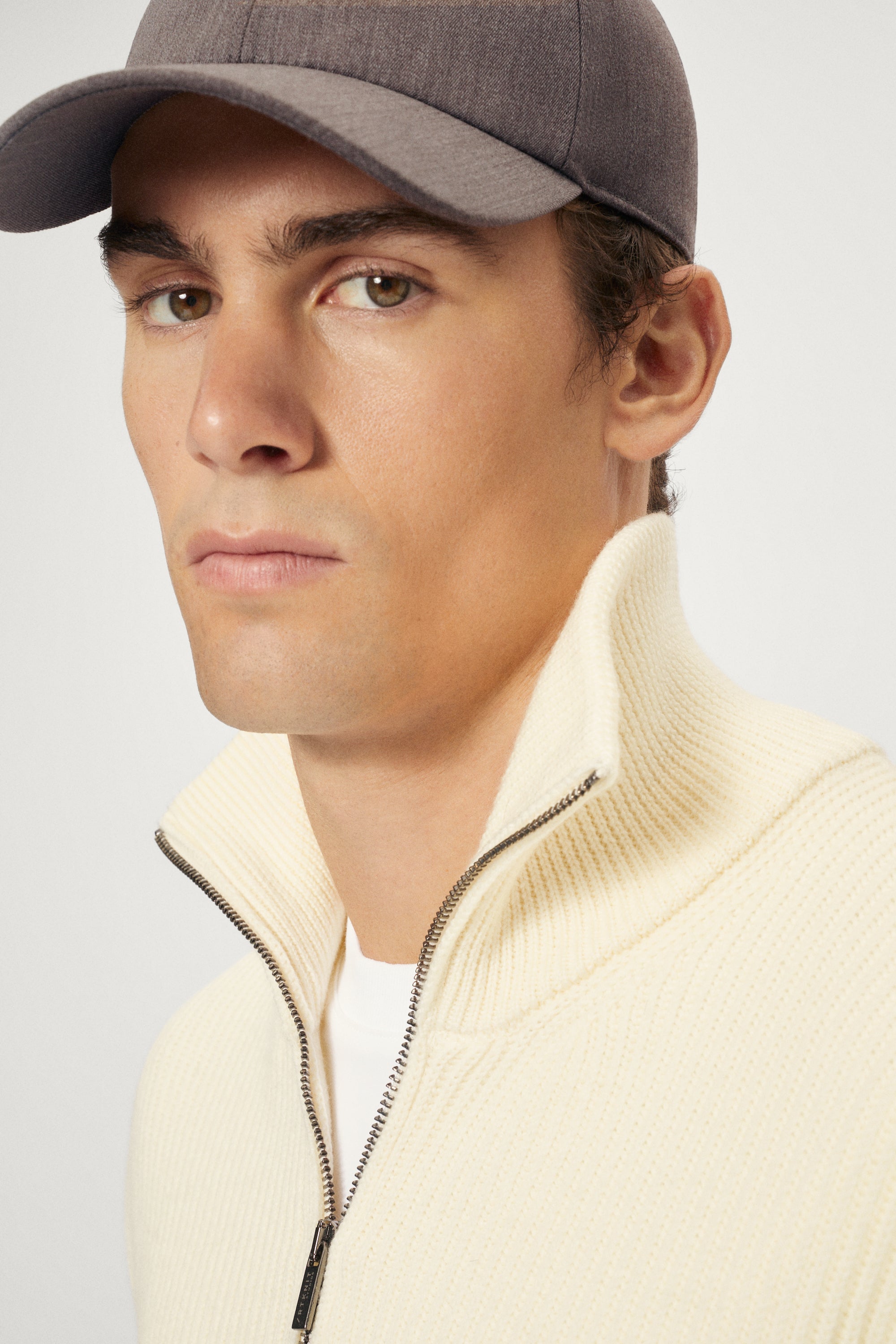 Man wearing a taupe hat and beige high-neck sweater against a light background
