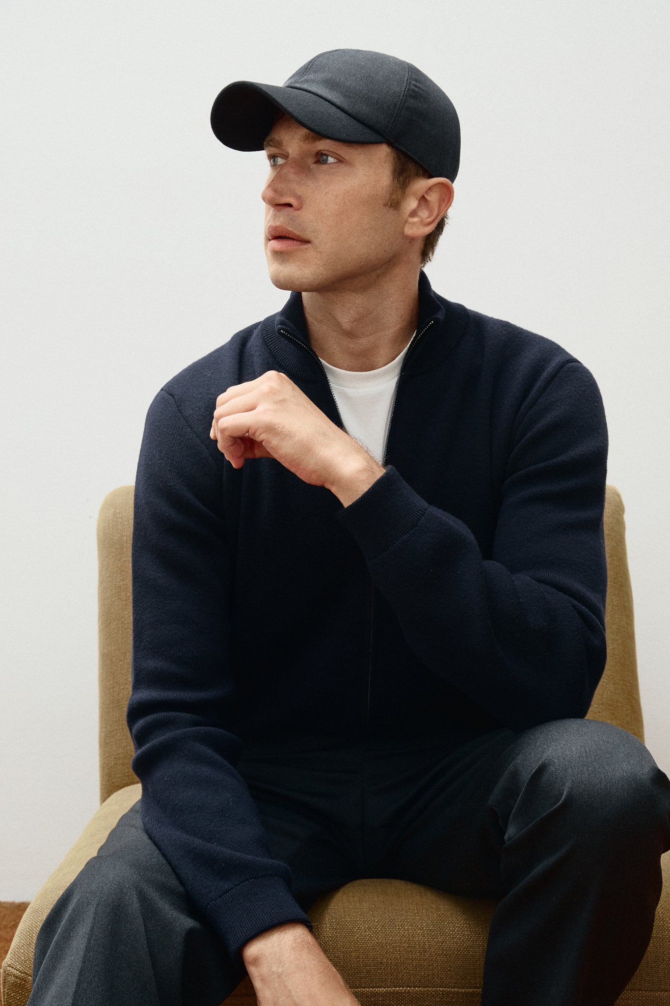Man wearing a navy sweater and hat sitting on a beige chair against a white wall