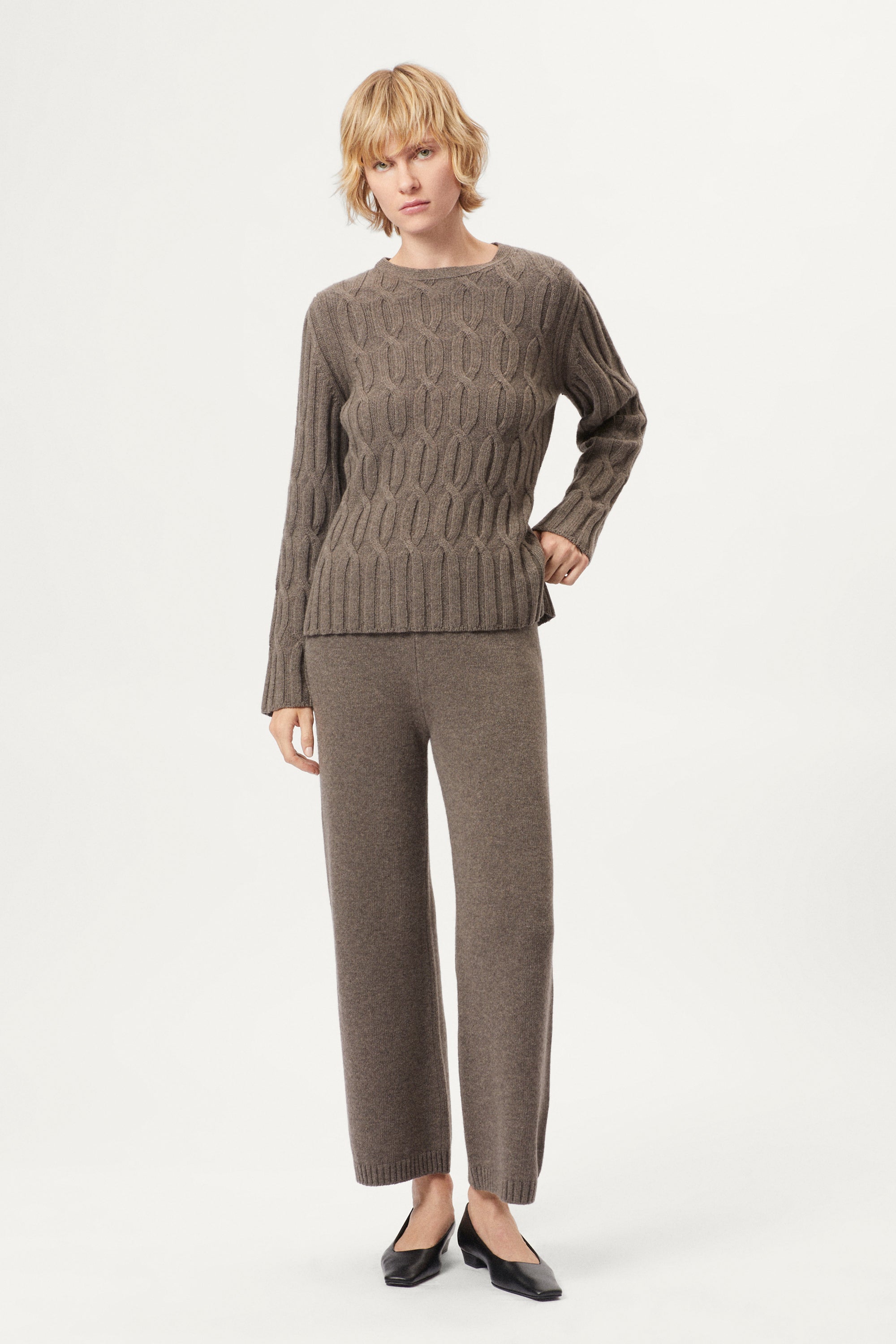 Woman wearing a woolen taupe cable knit sweater and pants on a white background