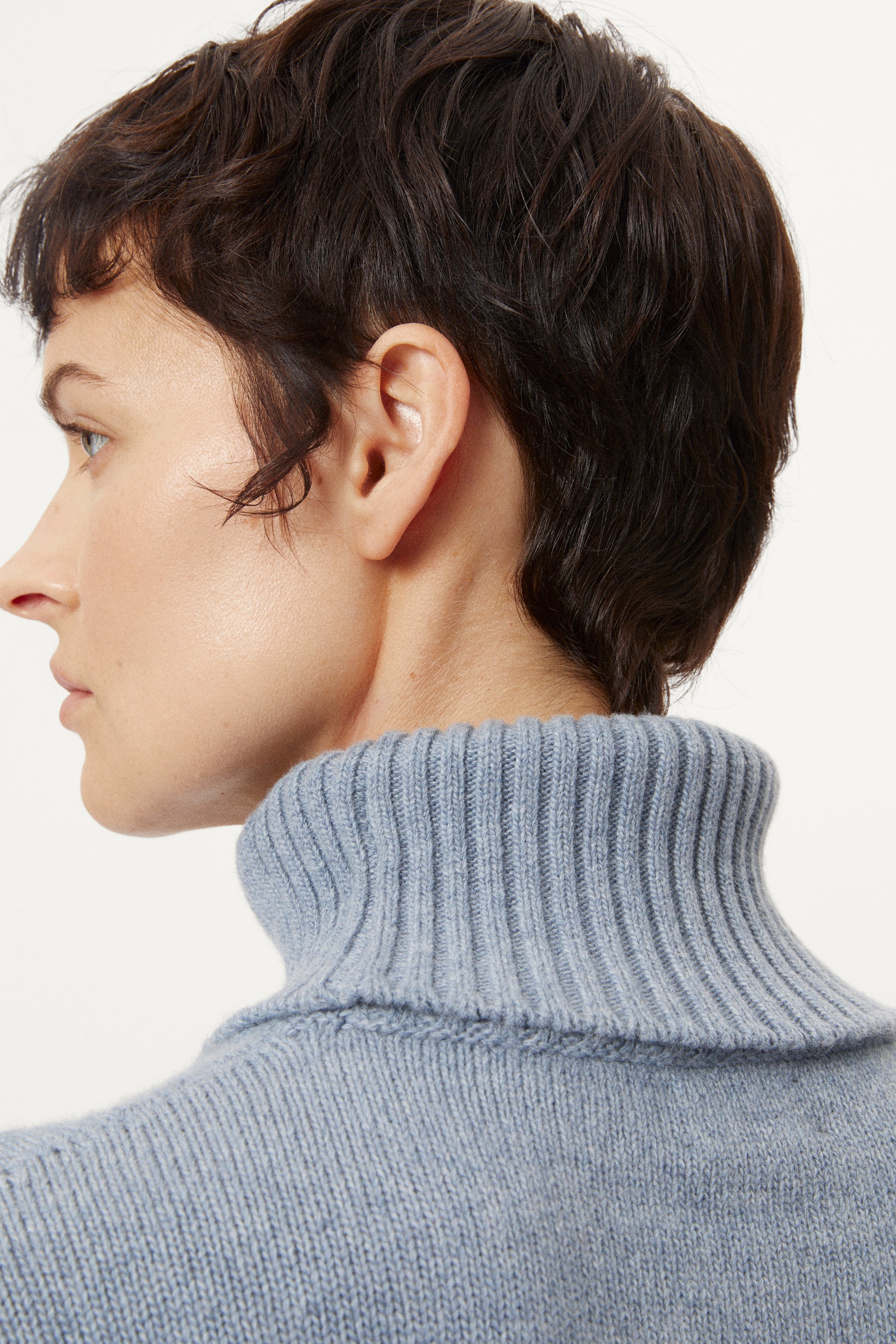 Person wearing a woolen light blue turtleneck sweater and dark pants on a white background