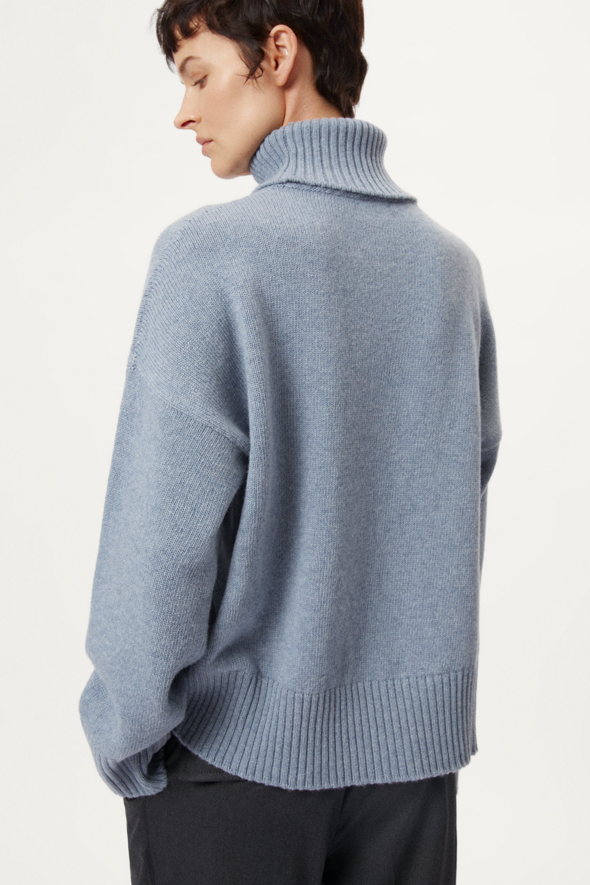 Person wearing a woolen light blue turtleneck sweater and dark pants on a white background