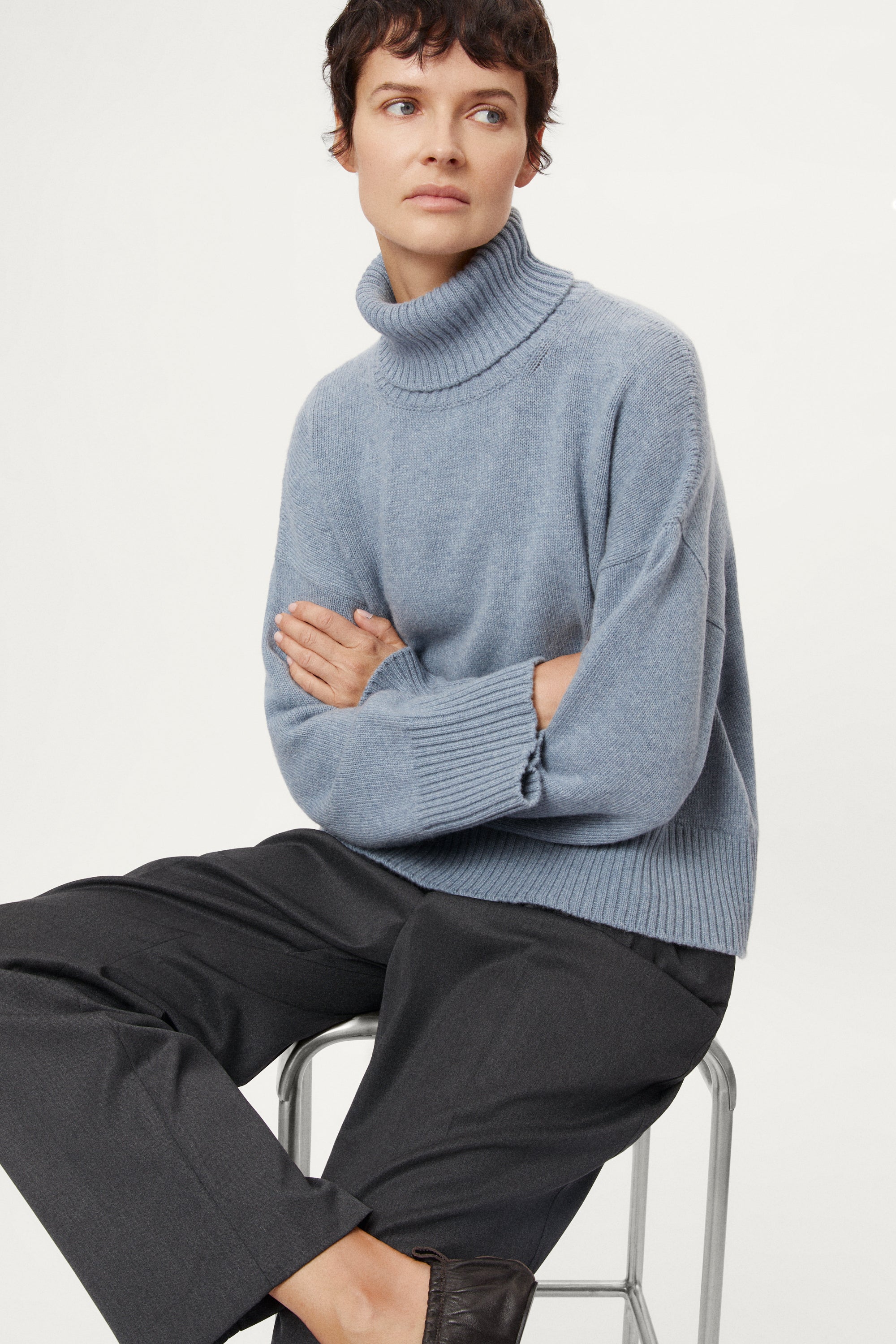 Person wearing a woolen light blue turtleneck sweater and dark pants on a white background