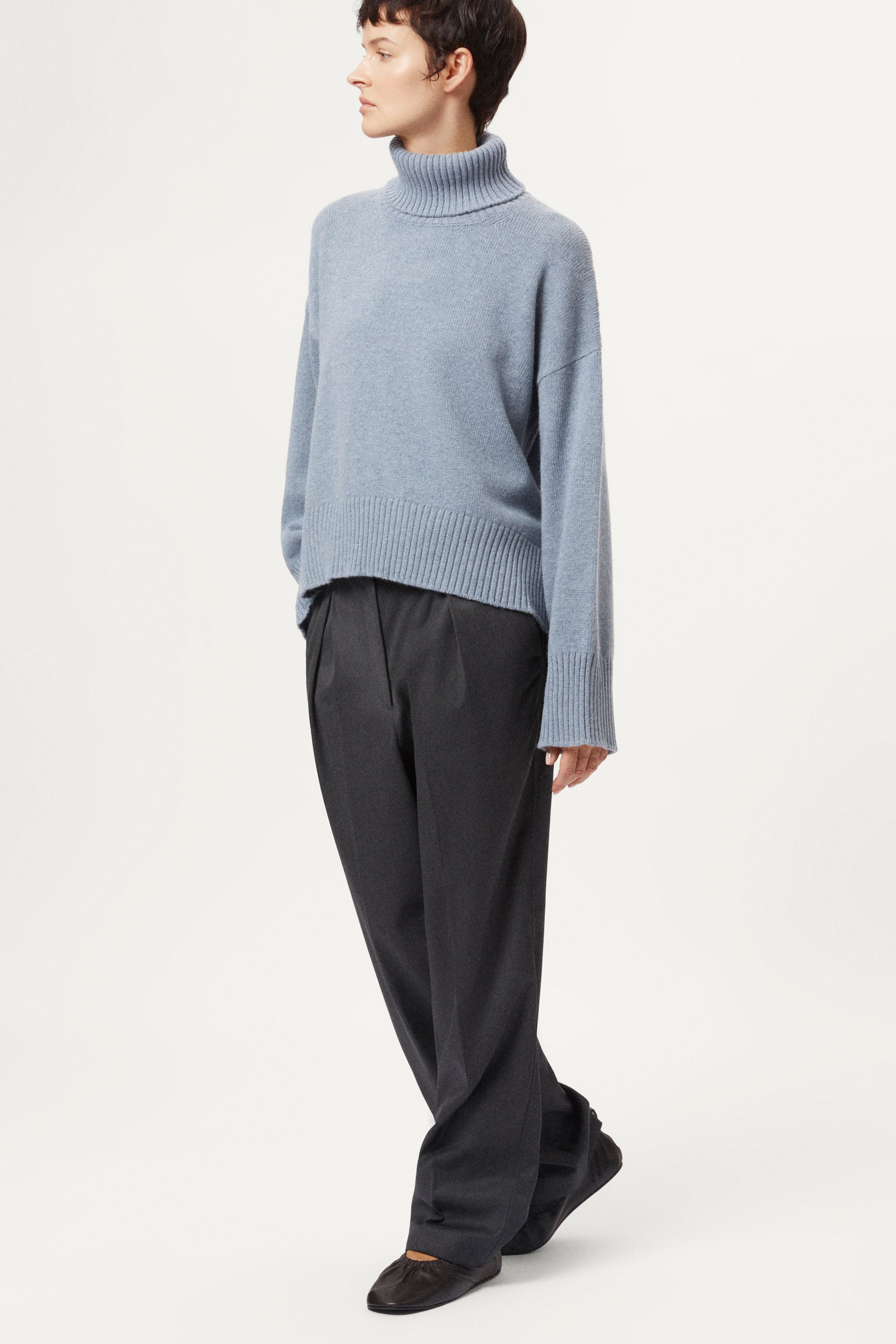Person wearing a woolen light blue turtleneck sweater and dark pants on a white background