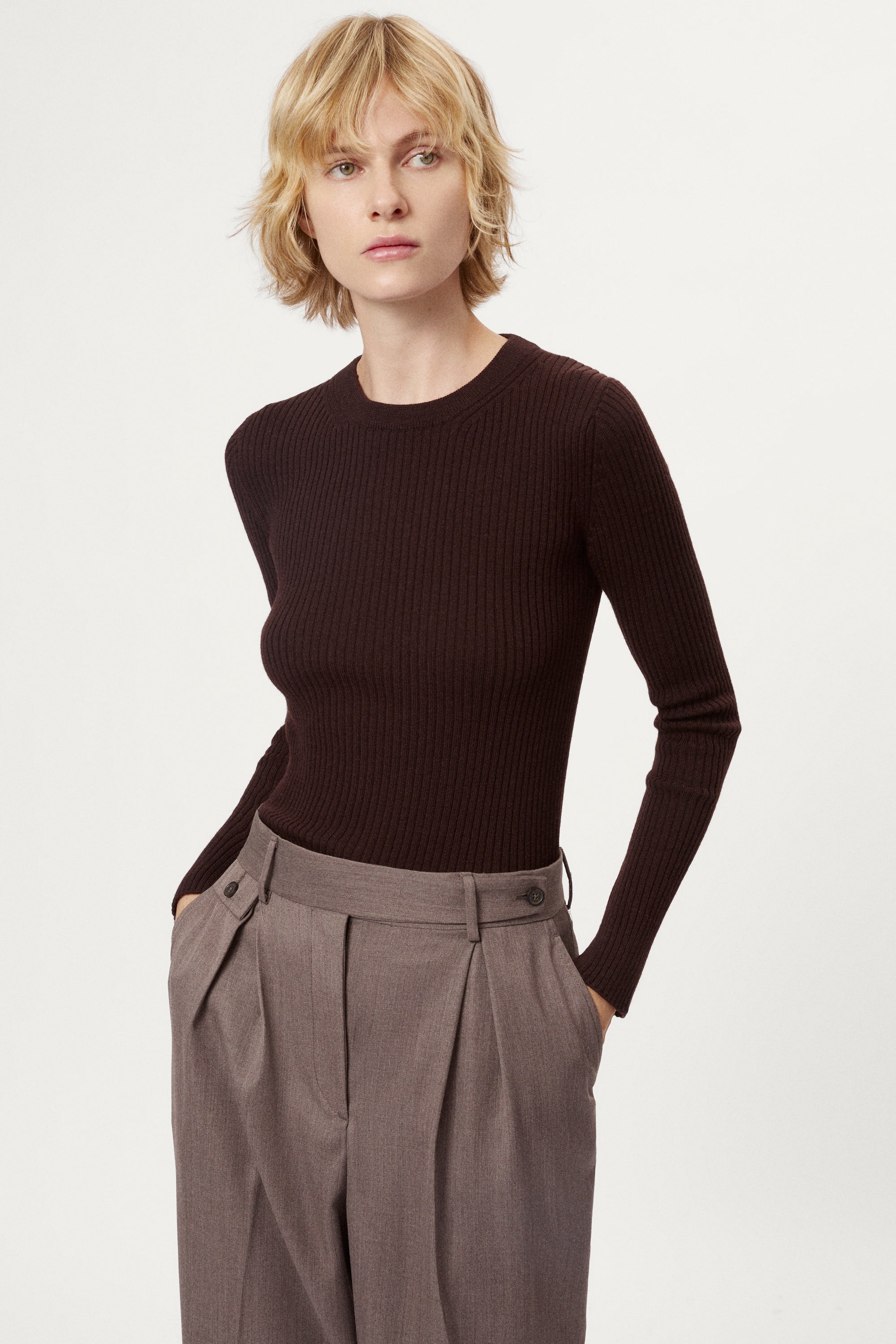 Woman wearing a dark brown sweater and light brown pleated pants on a white background