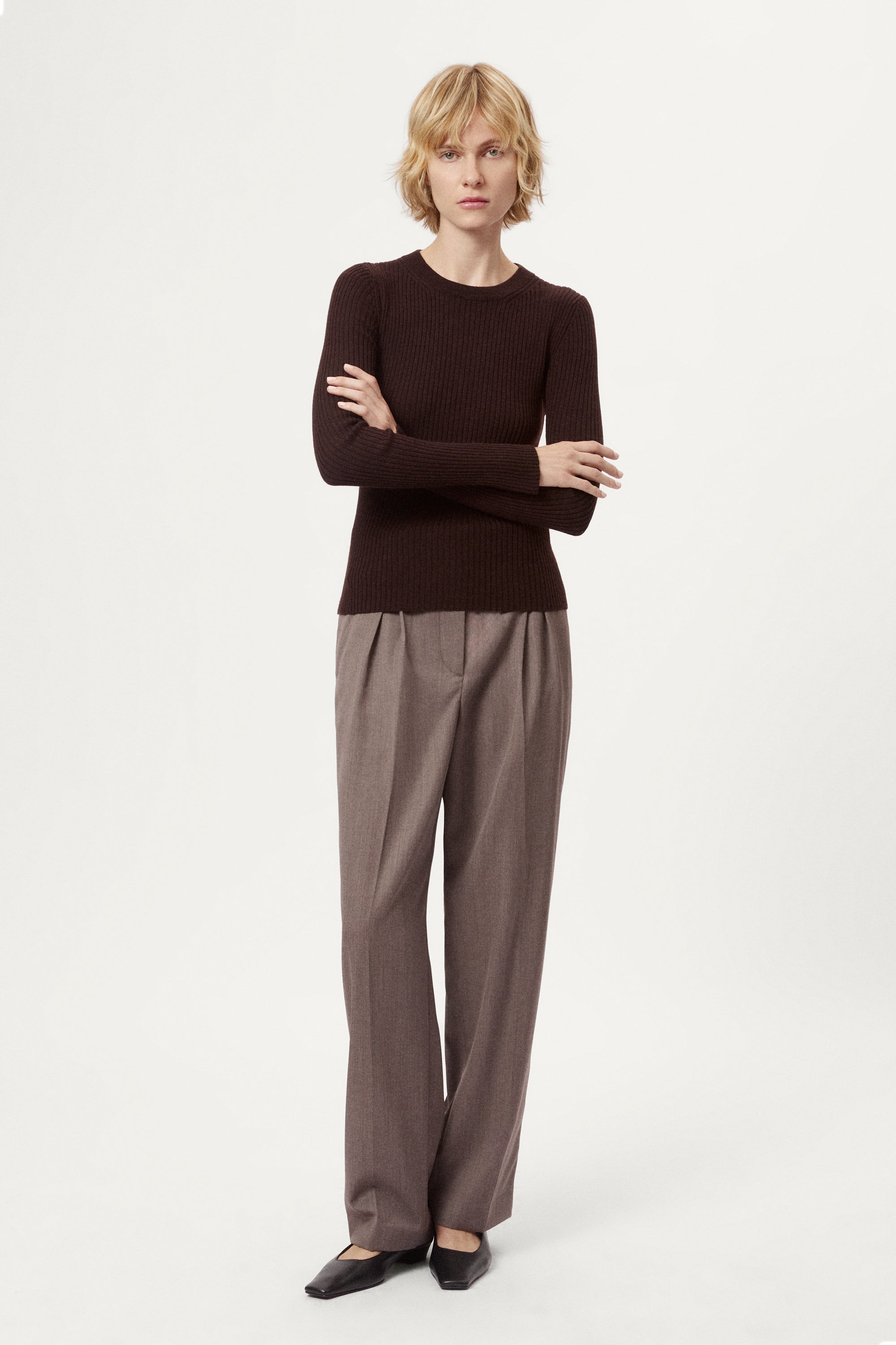 Woman wearing a dark brown sweater and light brown pleated pants on a white background
