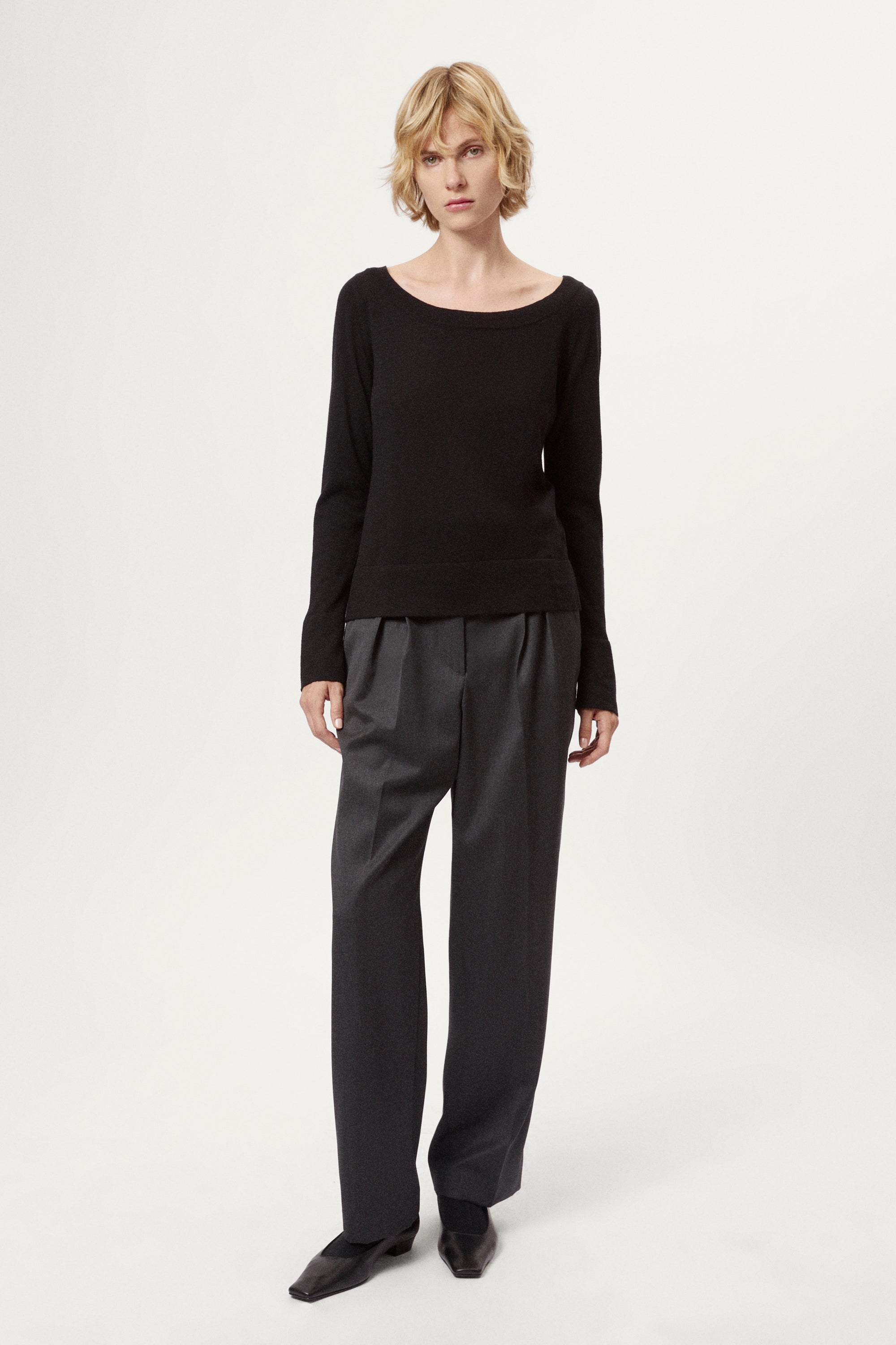 Person wearing a black long-sleeve top and dark gray pleated pants on a white background